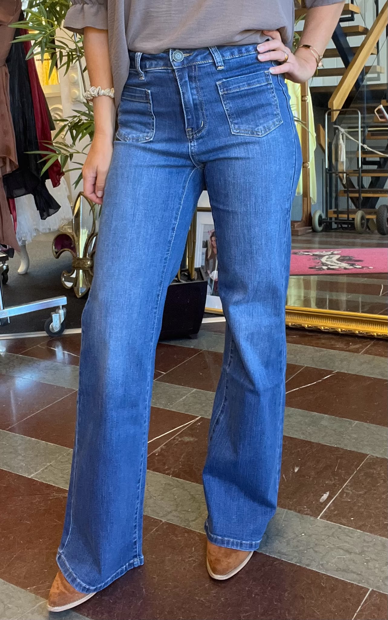 Person står med handen på höften och visar blå jeans med hög midja, stora framfickor och utsvängda ben. Fotograferad från midja till skor i en butiksmiljö med klädställning och spegel i bakgrunden.