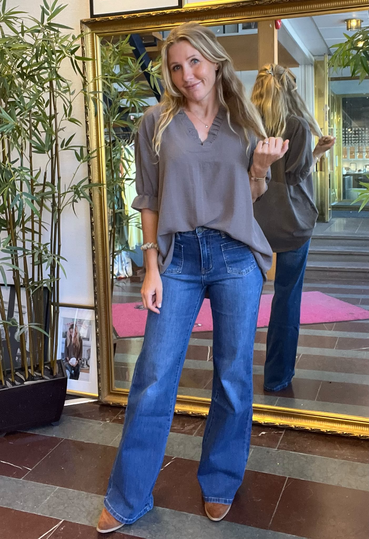 Kvinna med långt blont hår står och poserar framför en stor guldramsspegel, håller upp en hårslinga och ler. Hon bär grå blus, blå utsvängda jeans och bruna skor i en lobby med växter.
