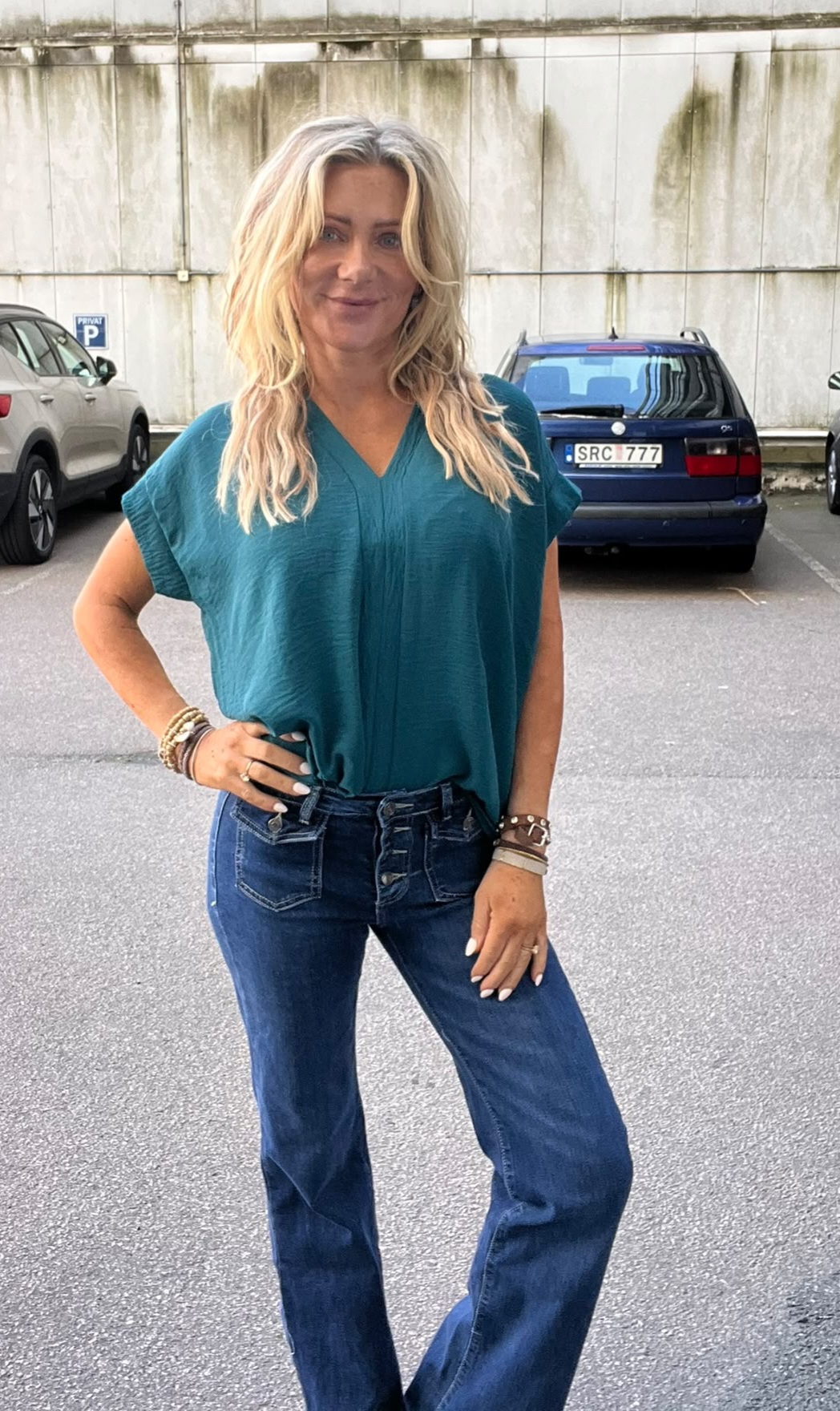 Leende kvinna med långt blont hår står och poserar med handen på höften på en parkeringsplats. Hon bär turkos blus, blå jeans och armband; bakom syns bilar och registreringsskylt “SRC 777”.