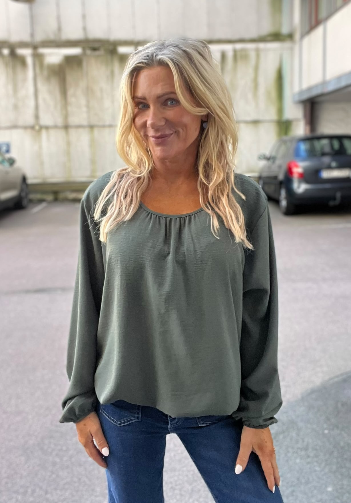 Blond kvinna står utomhus och ler lätt mot kameran, med händerna vid sidorna. Hon bär en mörkgrön långärmad blus och blå jeans. I bakgrunden syns en betongvägg och parkerade bilar.
