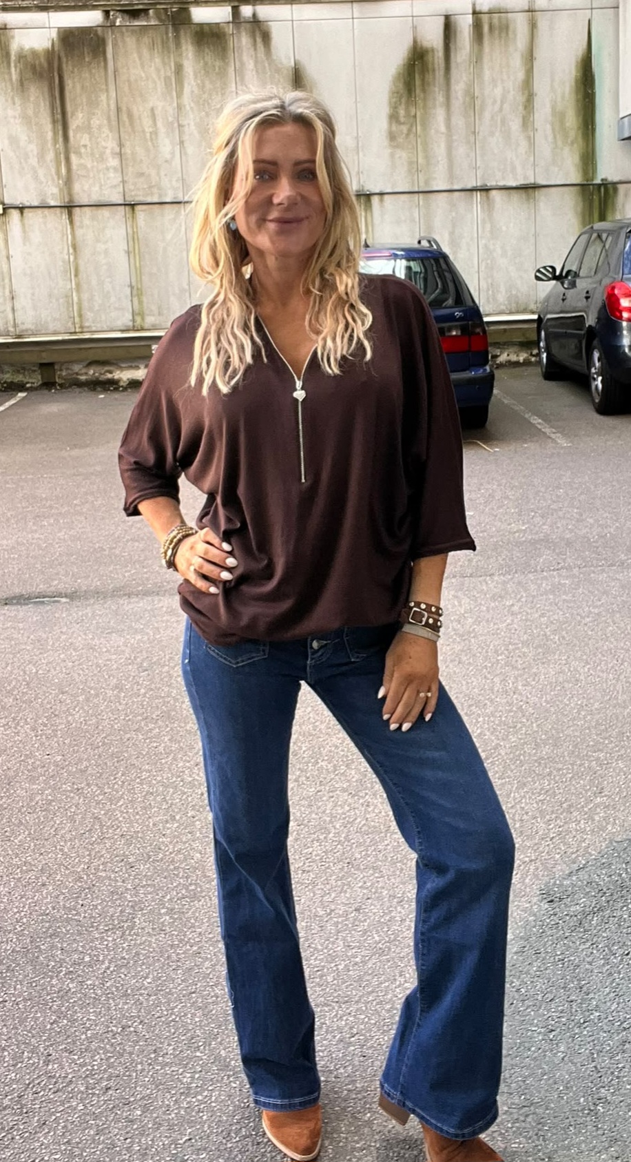 Kvinna med långt blont hår poserar med handen på höften och ler mot kameran. Hon bär mörkbrun topp med dragkedja, blå utsvängda jeans och bruna skor, på en parkeringsplats framför en betongvägg och bilar.