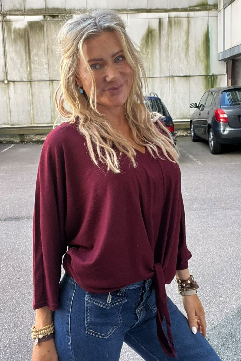 Blond kvinna ler mot kameran och poserar med ena benet böjt. Hon bär vinröd, lös blus och blå jeans med armband och örhängen. Bakom syns parkerade bilar och en betongvägg med gröna fläckar.