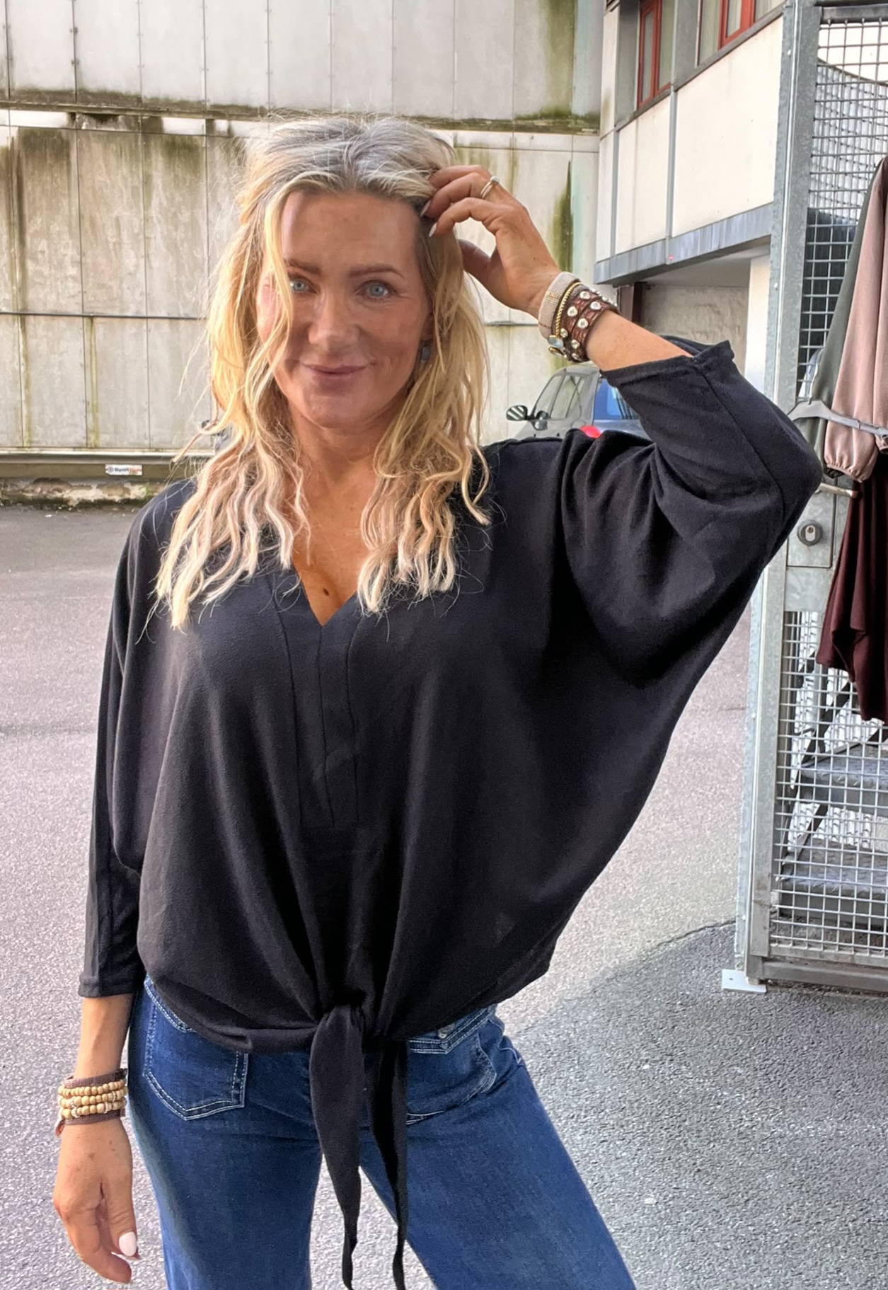 Leende kvinna med långt blont hår står utomhus vid en byggnad och en grind, lyfter handen mot håret och tittar mot kameran. Hon bär svart blus med knut framtill, jeans och flera armband.
