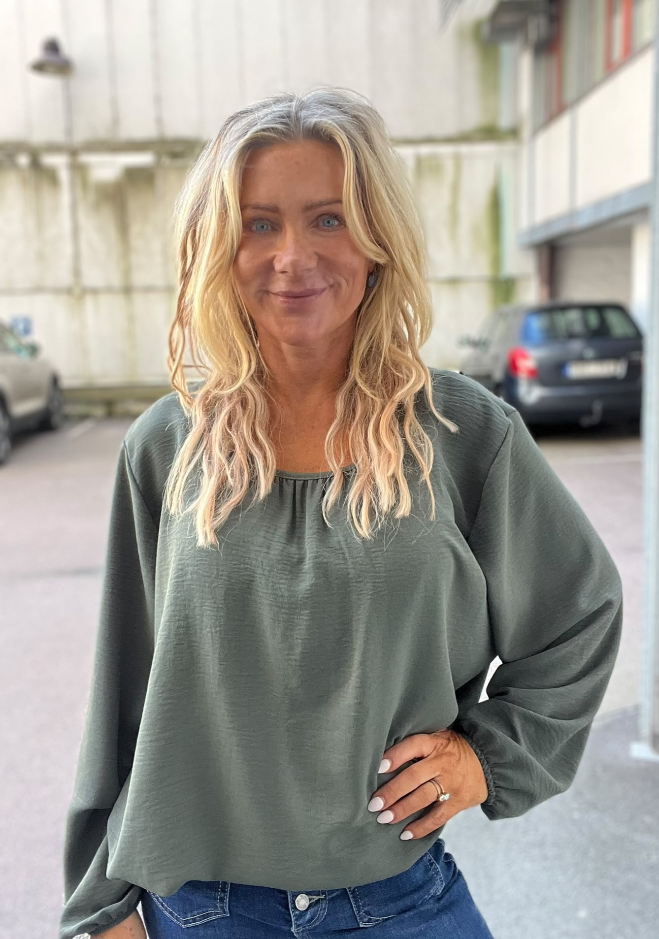 Leende kvinna med långt, vågigt blont hår står och tittar mot kameran med handen på höften. Hon bär en grön, långärmad blus och jeans. Bakgrunden visar en gård med betongväggar och parkerade bilar.