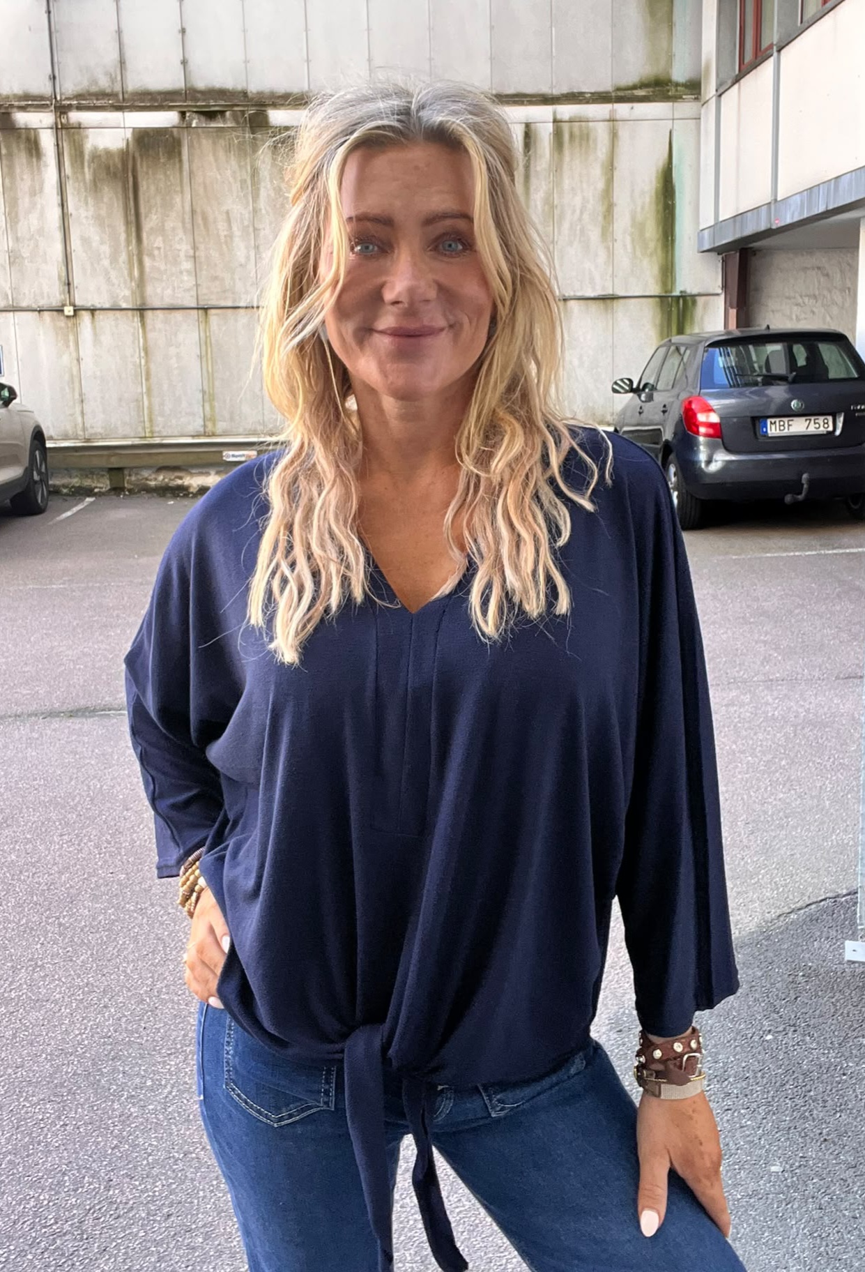 Kvinna med långt blont hår står och ler mot kameran, med ena handen på höften. Hon bär mörkblå blus och jeans. I bakgrunden syns en parkeringsplats vid en betongvägg och en bil med registreringsskylt “MBF 758”.