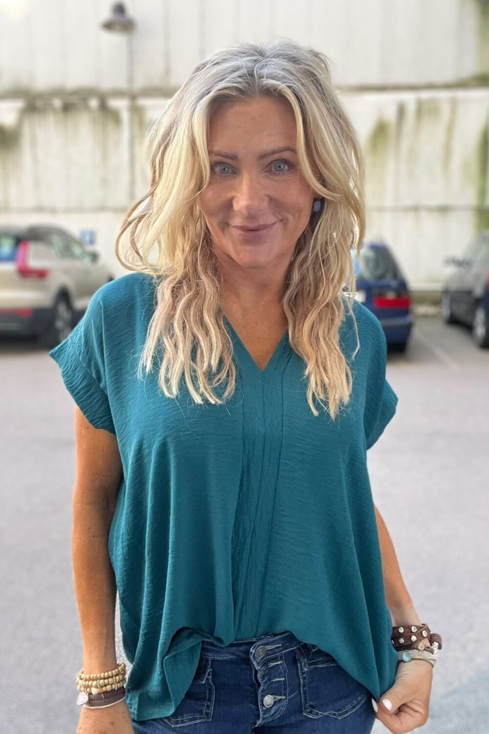 Leende kvinna med långt blont hår står och tittar mot kameran, klädd i turkos blus och jeans, med armband på handlederna. Hon står utomhus på en parkering med bilar och ljus vägg i bakgrunden.