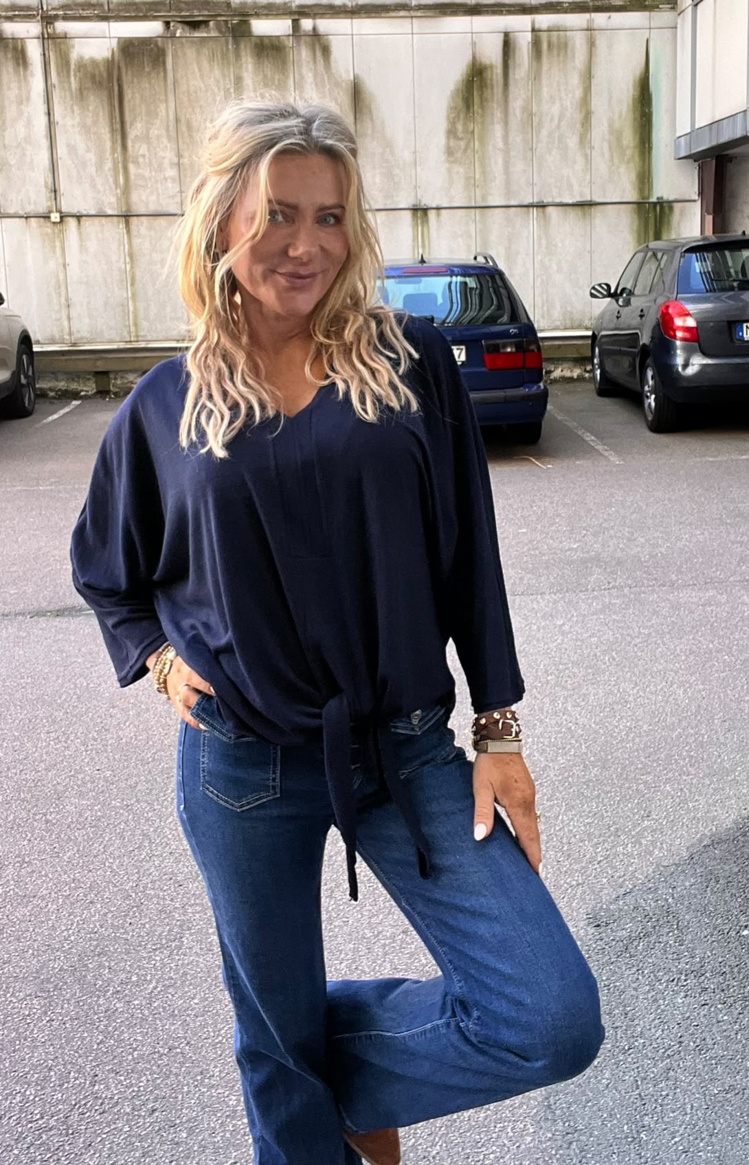 Kvinna med långt blont hår poserar leende på en parkeringsplats, med ena handen i jeansfickan och ena benet böjt. Hon bär mörkblå knytblus, vida jeans och armband; bilar och betongvägg i bakgrunden.