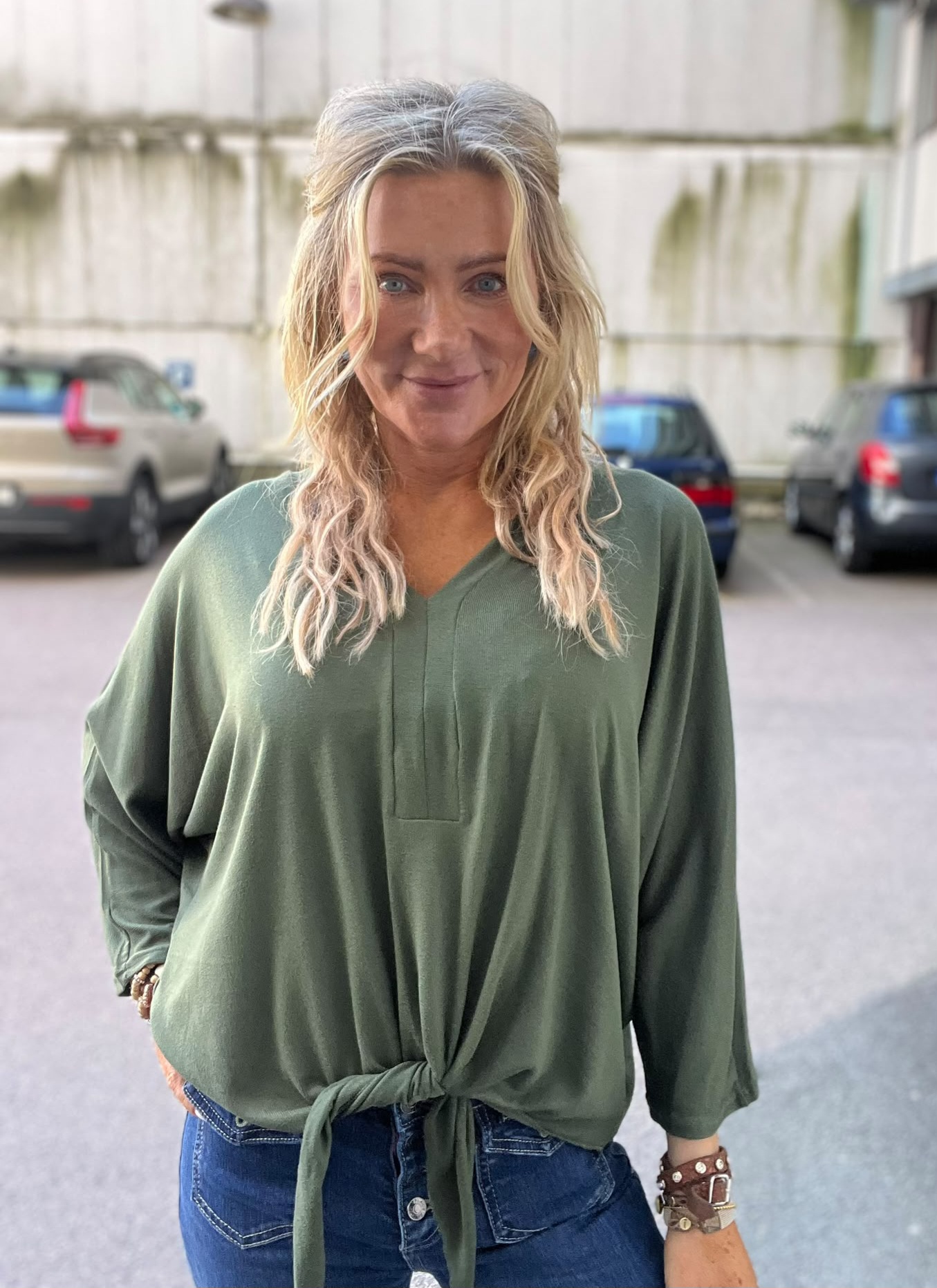 Kvinna med långt blont hår står och ler mot kameran, iklädd grön knuten blus och blå jeans med armband. Hon befinner sig utomhus på en parkeringsplats med bilar och en ljus vägg i bakgrunden.