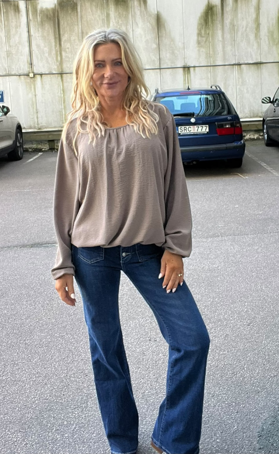 Kvinna med långt blont hår står och poserar med ett ben fram, iklädd grå blus och blå jeans, på en parkeringsplats. Bakom henne syns bilar och en betongvägg; registreringsskylt: “SRC 777”.