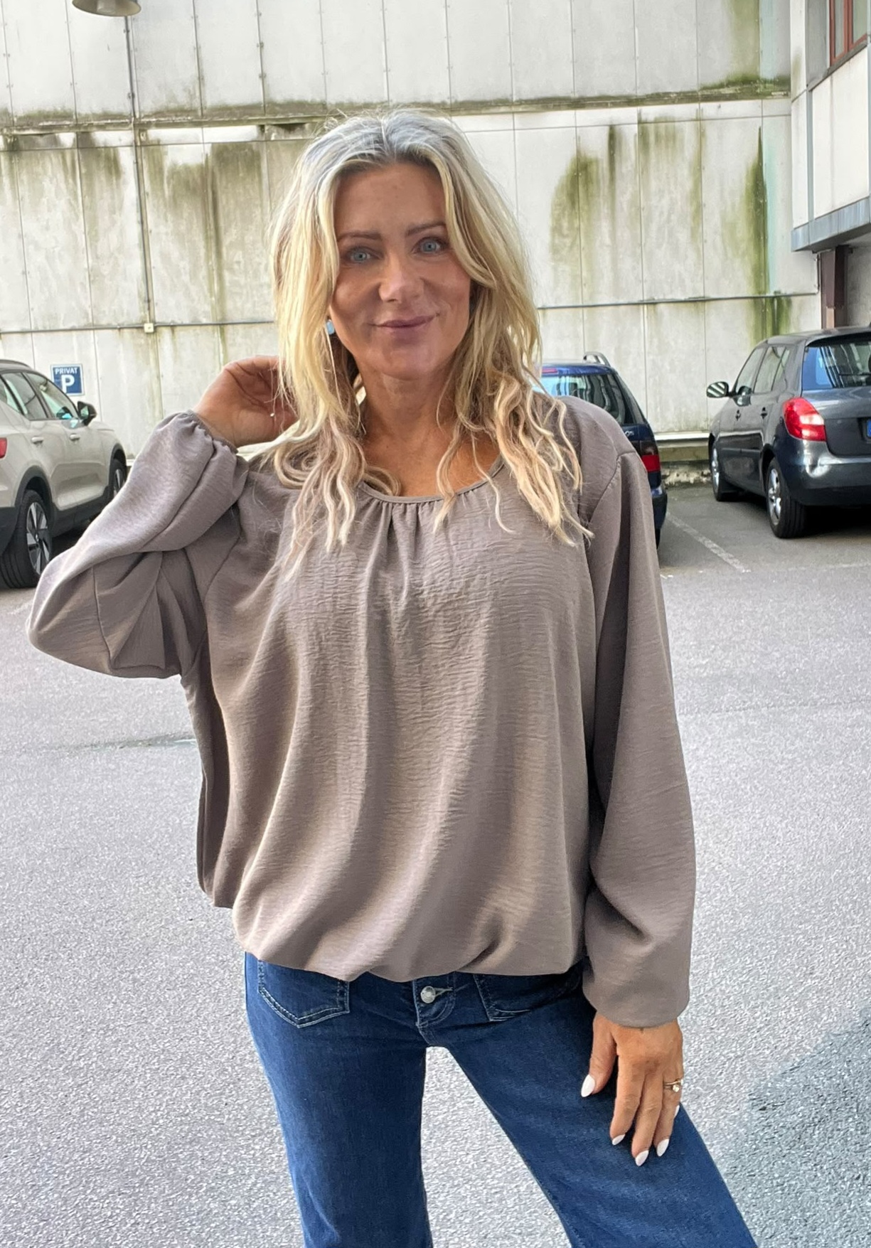 Leende kvinna med långt blont hår står i en parkeringsmiljö och rör vid håret med ena handen. Hon bär en lös beige blus med vida ärmar och blå jeans; bilar och betongvägg syns bakom.