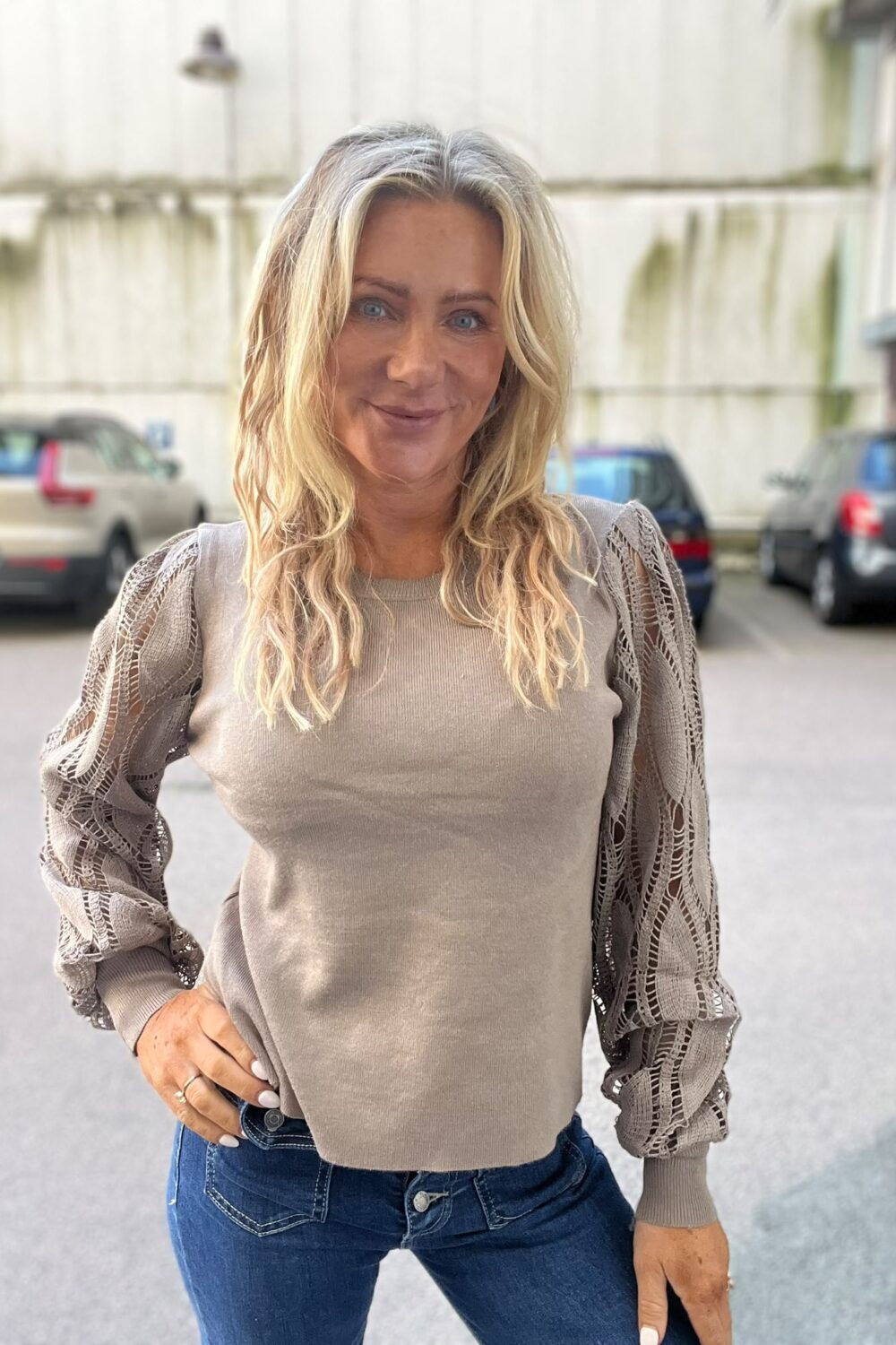 Kvinna med långt blont hår står och ler mot kameran med ena handen i jeansfickan. Hon bär en beige stickad tröja med spetsiga, volangärmar. Bakgrunden visar en parkeringsplats med bilar och en ljus byggnad.