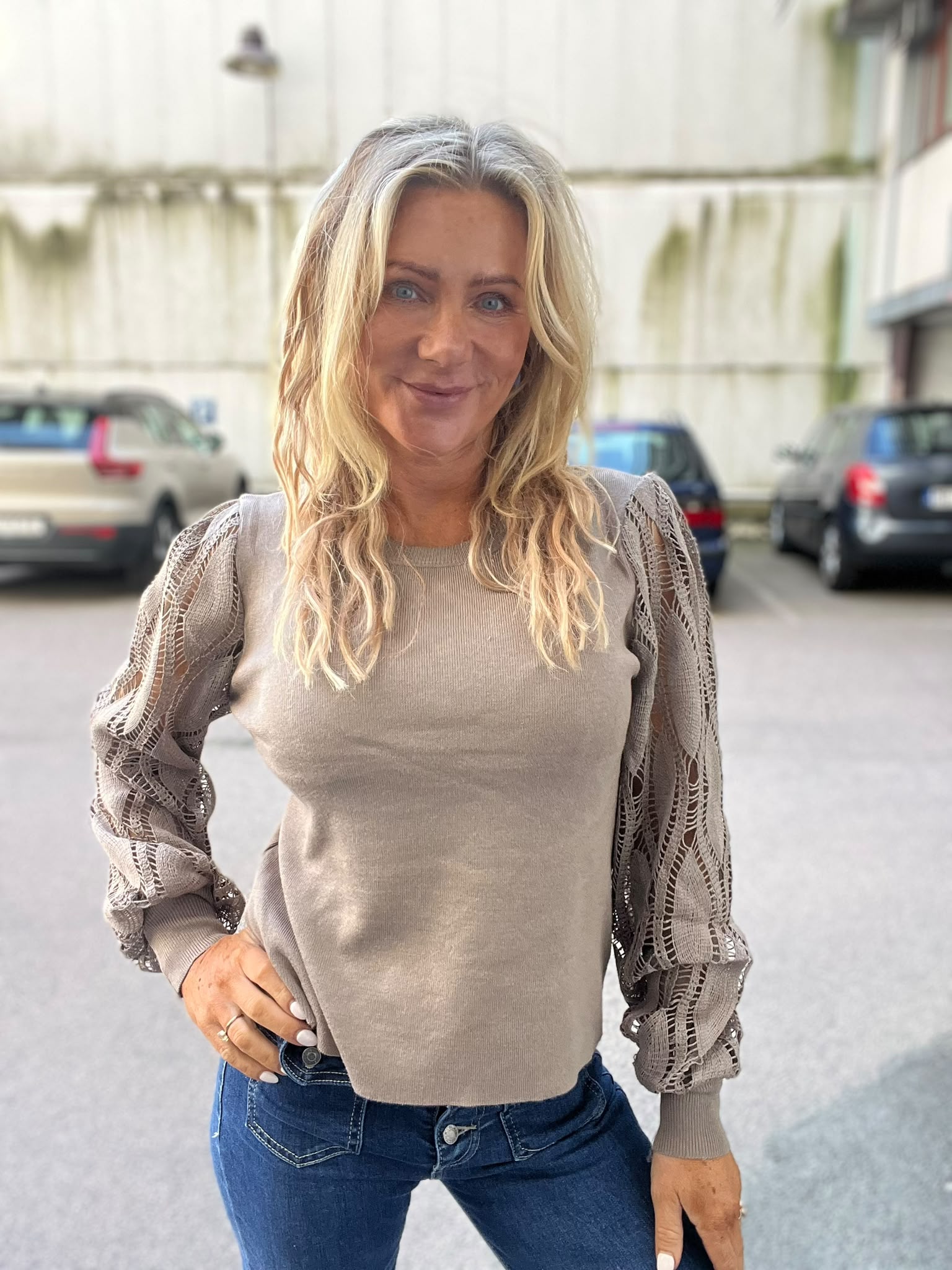 Kvinna med långt blont hår står och ler mot kameran med ena handen i jeansfickan. Hon bär en beige stickad tröja med spetsiga, volangärmar. Bakgrunden visar en parkeringsplats med bilar och en ljus byggnad.