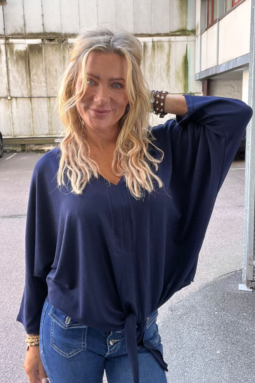 Leende kvinna med långt blont hår poserar med ena armen bakom huvudet och tittar mot kameran. Hon bär en mörkblå blus och jeans med armband, stående utomhus vid en industribyggnad och parkering.