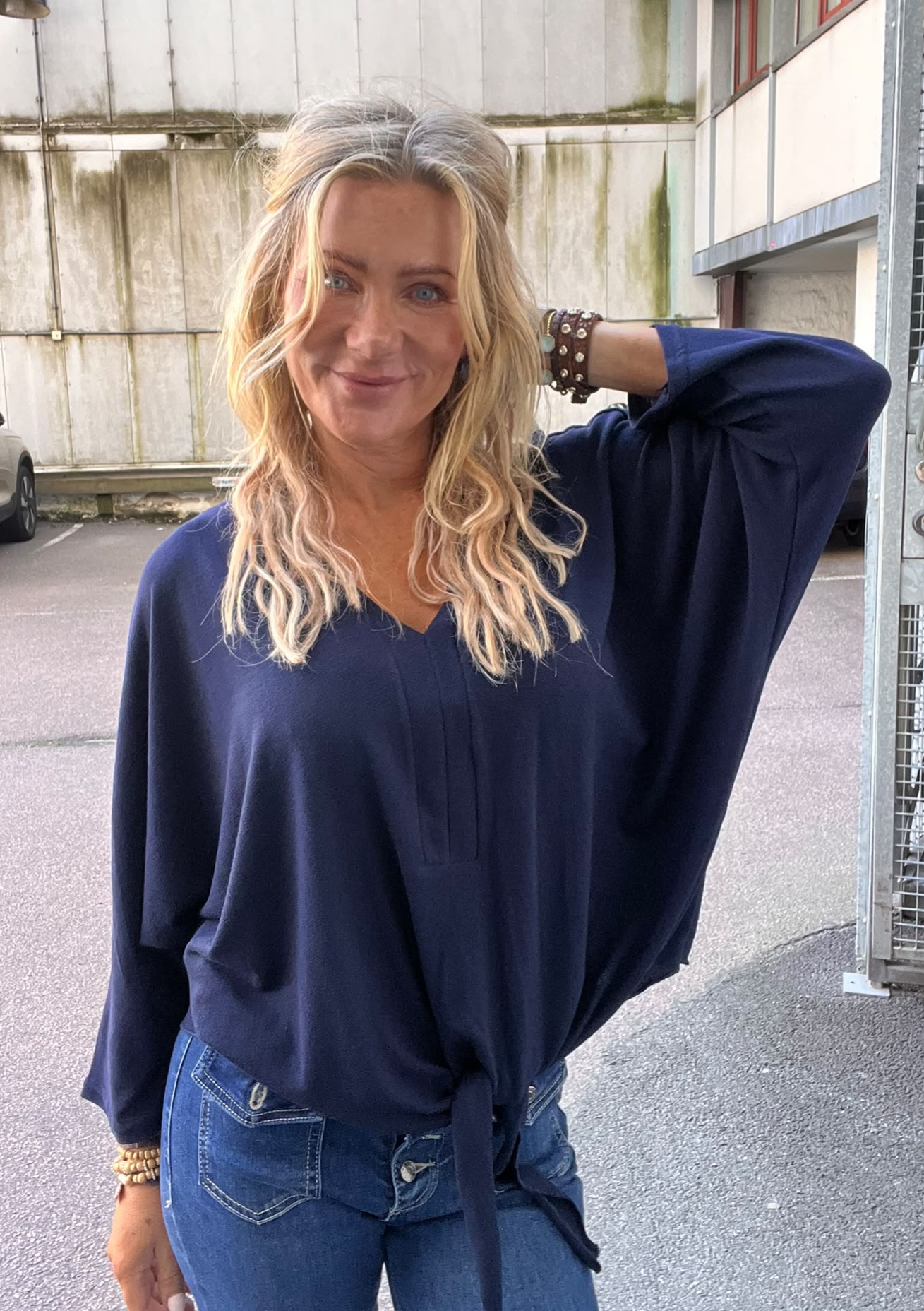 Leende kvinna med långt blont hår poserar med ena armen bakom huvudet och tittar mot kameran. Hon bär en mörkblå blus och jeans med armband, stående utomhus vid en industribyggnad och parkering.