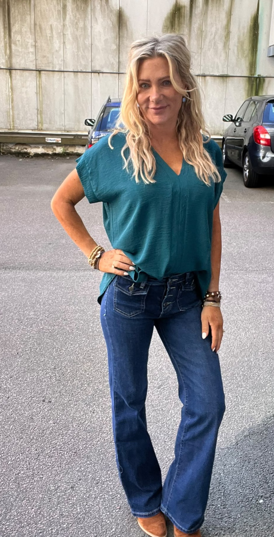 Blond kvinna poserar med handen på höften och ler mot kameran. Hon bär turkos V-ringad blus, blå bootcut-jeans och armband. Fotot är taget på en parkeringsplats med bilar och en betongvägg i bakgrunden.