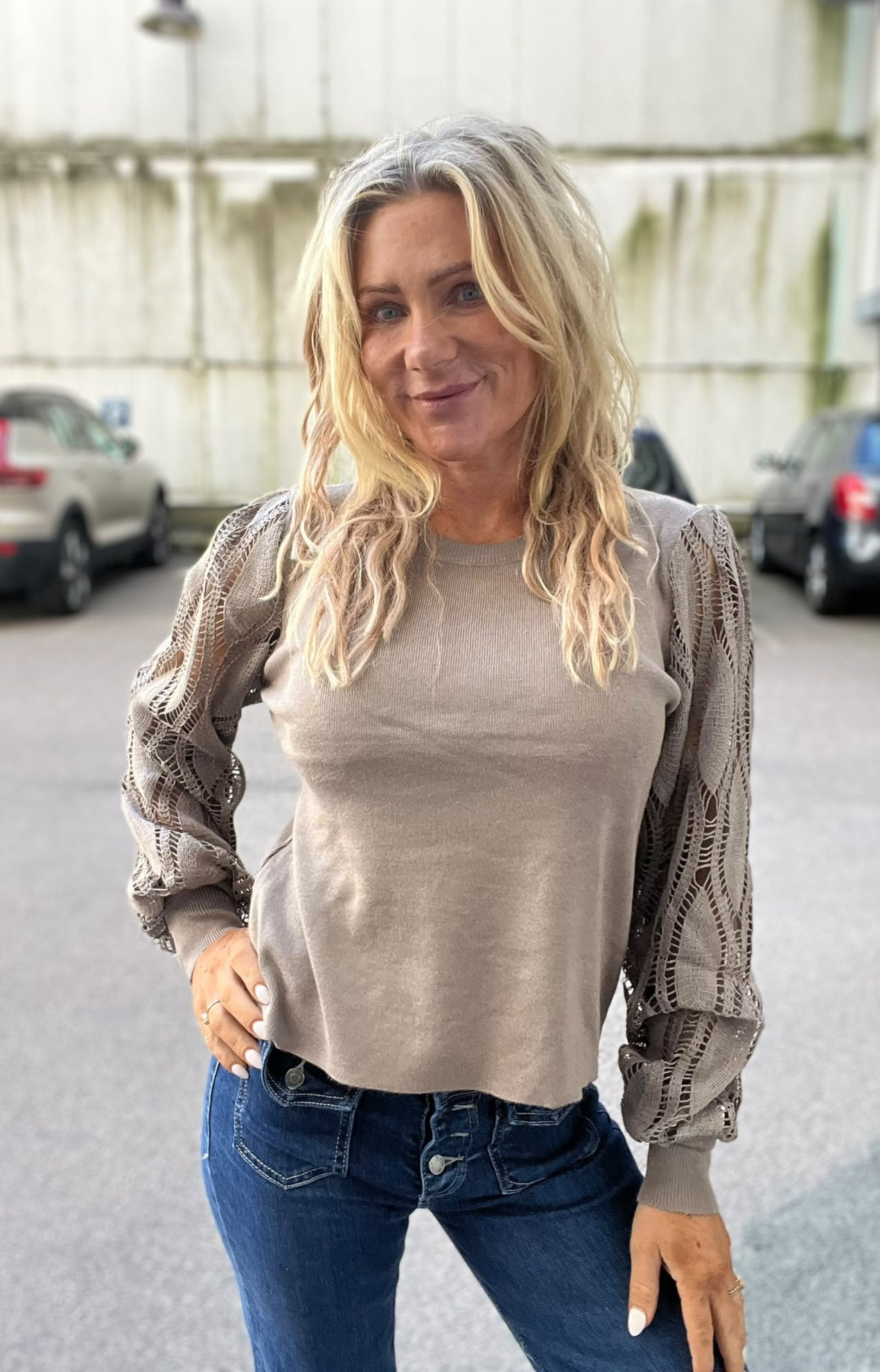 Blond kvinna står och poserar med en hand i jeansfickan och ler mot kameran. Hon bär en beige stickad tröja med spetsmönstrade ärmar och blå jeans, med parkerade bilar och en ljus byggnad i bakgrunden.