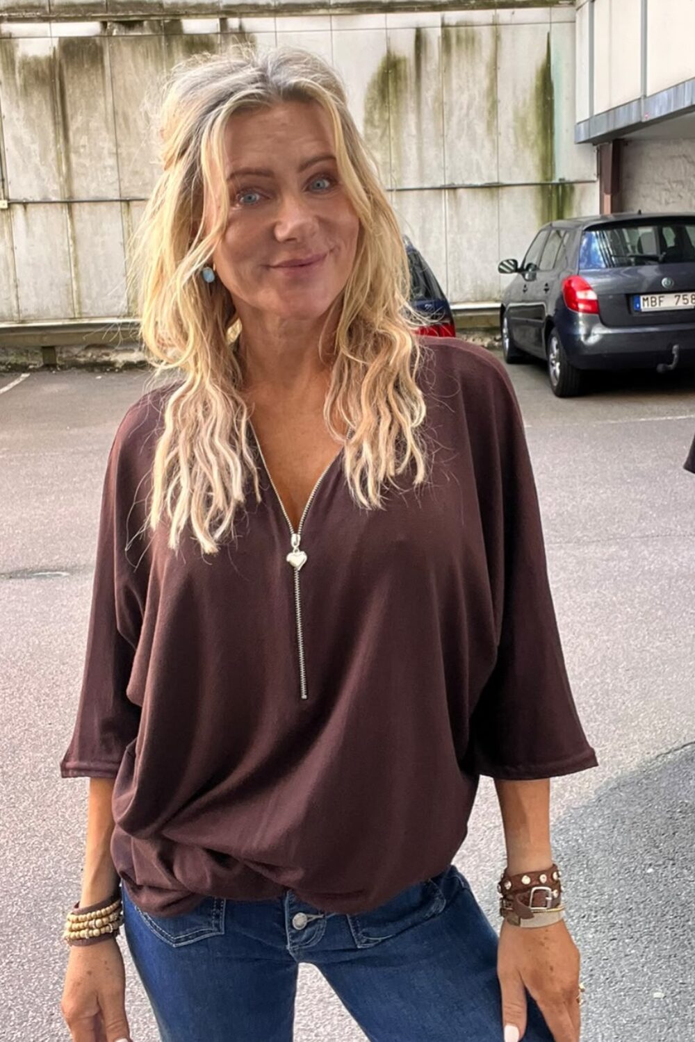 Kvinna med långt blont hår ler mot kameran och står avslappnat med händerna vid sidorna. Hon bär mörk topp med dragkedja och jeans, på en parkeringsplats med bilar och betongvägg i bakgrunden.