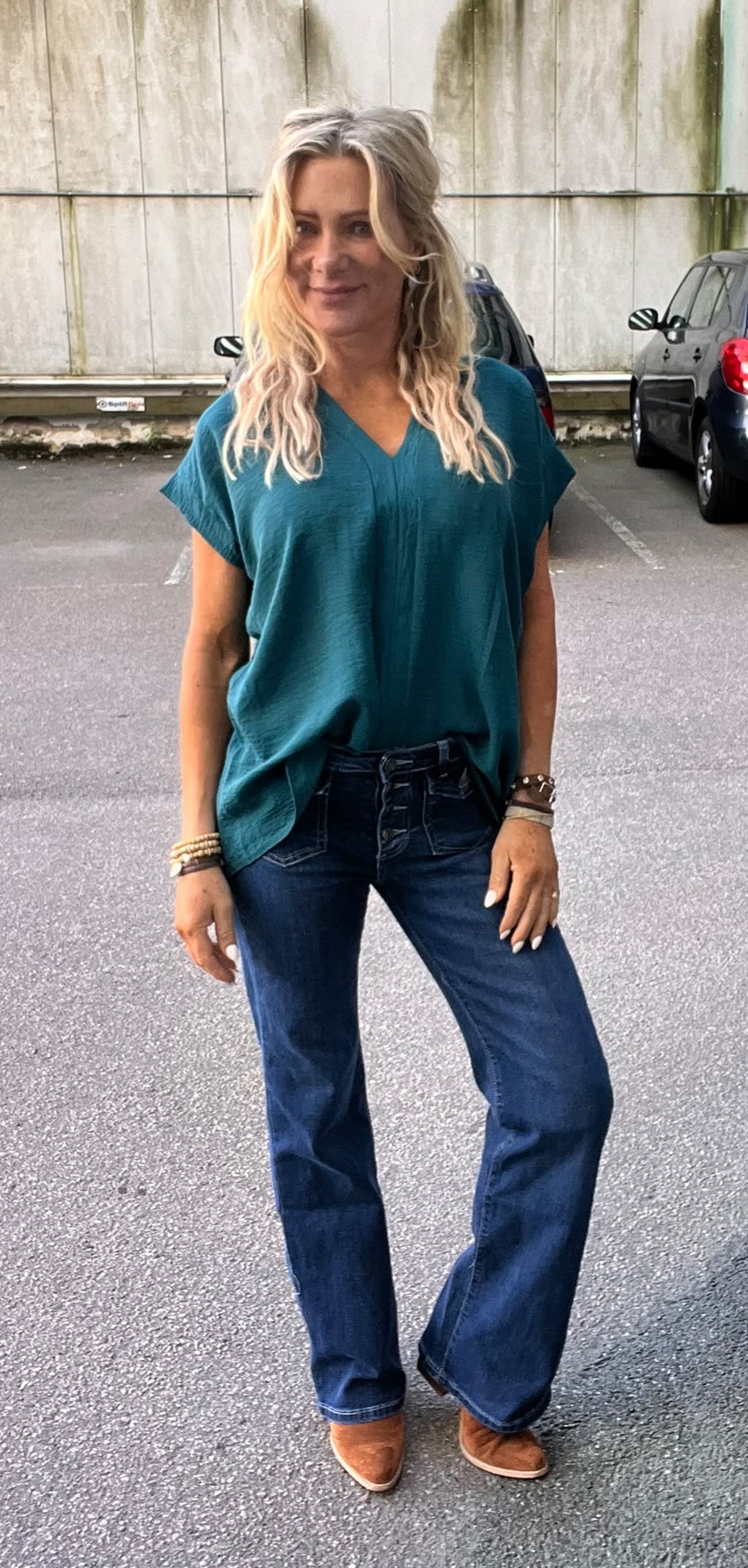 Blond kvinna står och ler mot kameran på en parkeringsplats, klädd i turkos V-ringad topp och utsvängda blå jeans med knappar, bruna skor och flera armband. Bilar och betongvägg syns i bakgrunden.