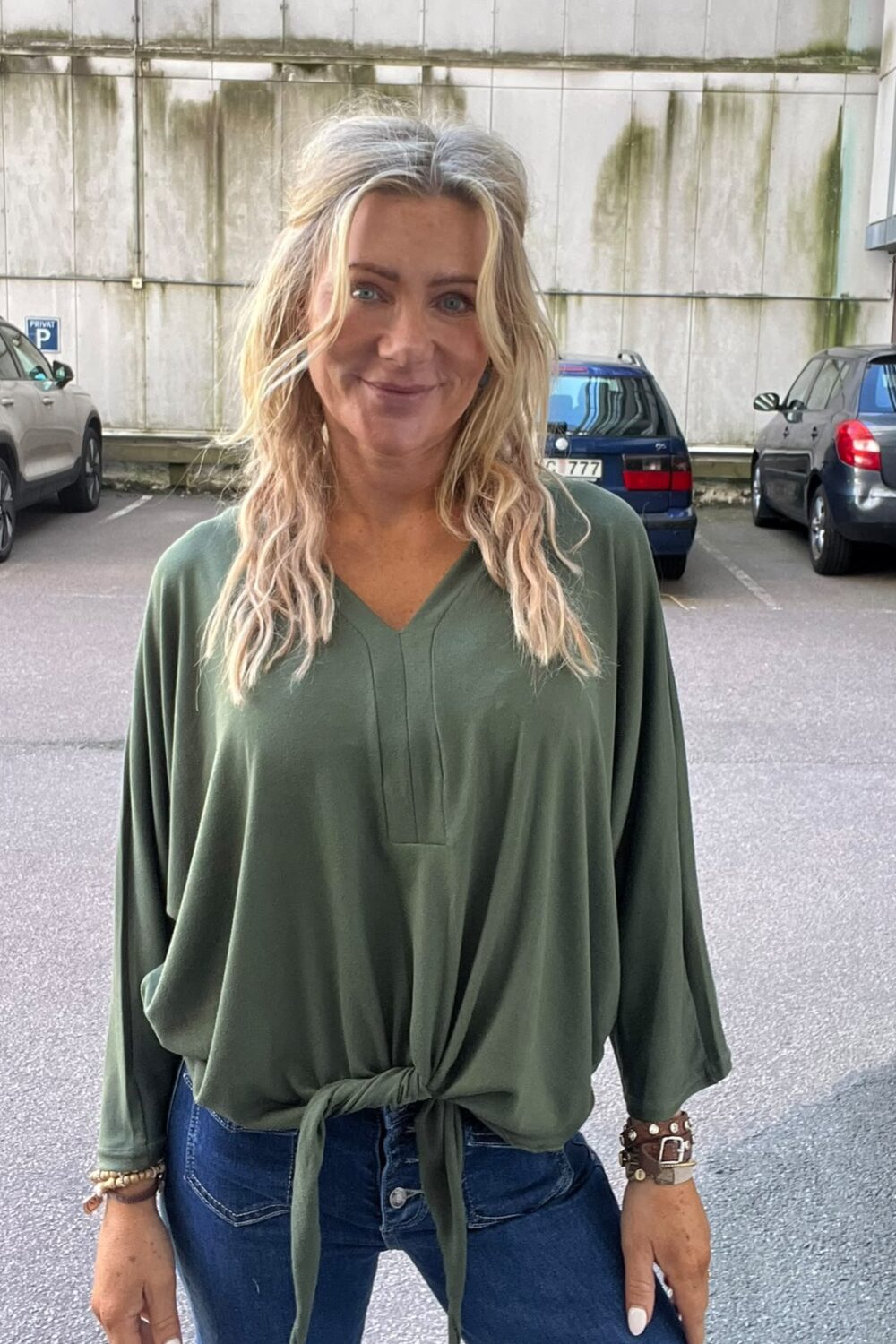 Blond kvinna med vågigt hår står och ler mot kameran på en parkeringsplats. Hon bär en grön, lös blus knuten framtill och blå jeans, med armband. Bakom syns parkerade bilar och en sliten betongvägg.