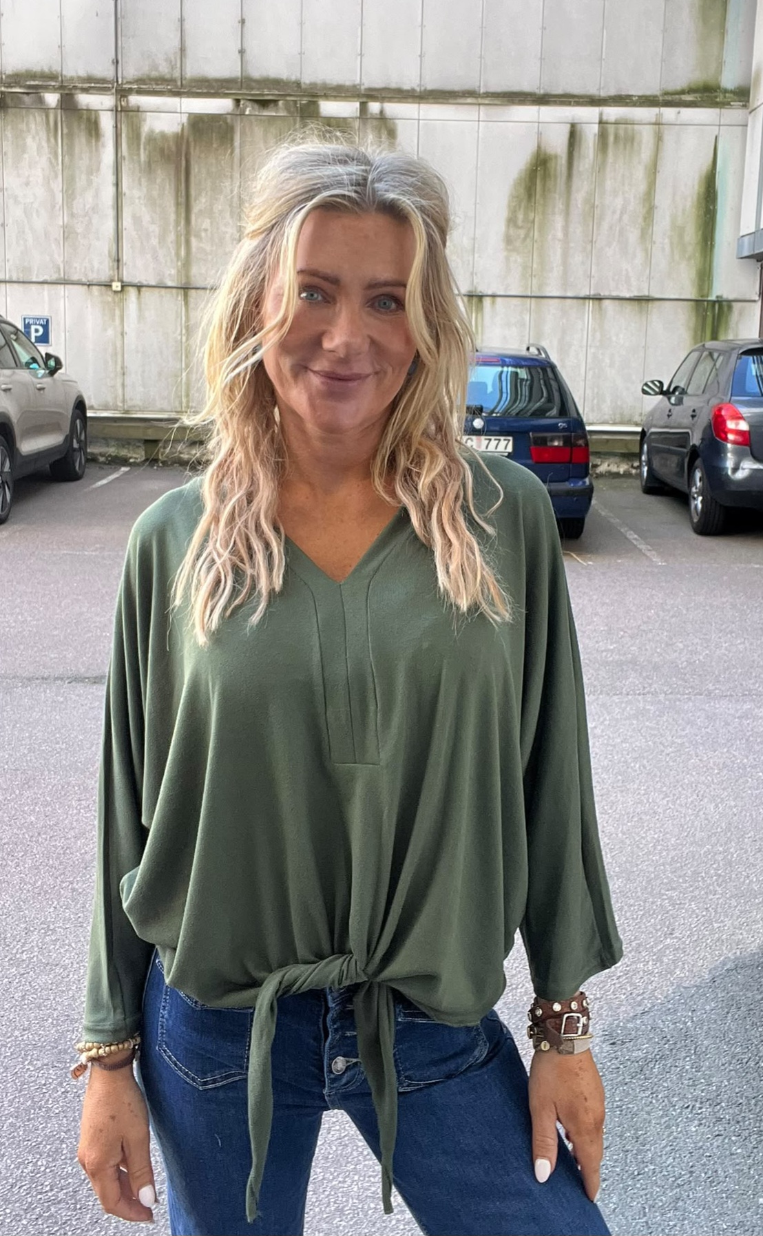 Blond kvinna med vågigt hår står och ler mot kameran på en parkeringsplats. Hon bär en grön, lös blus knuten framtill och blå jeans, med armband. Bakom syns parkerade bilar och en sliten betongvägg.