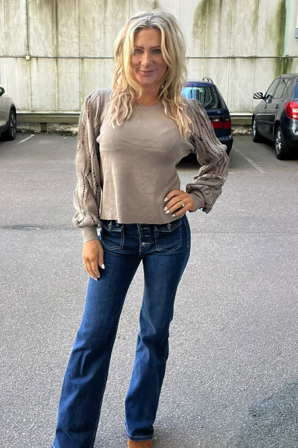 Blond kvinna står och poserar med ena handen på höften på en parkeringsplats. Hon bär beige topp med spetsiga ballongärmar, mörkblå bootcut-jeans och bruna skor; bilar och en betongvägg syns i bakgrunden.