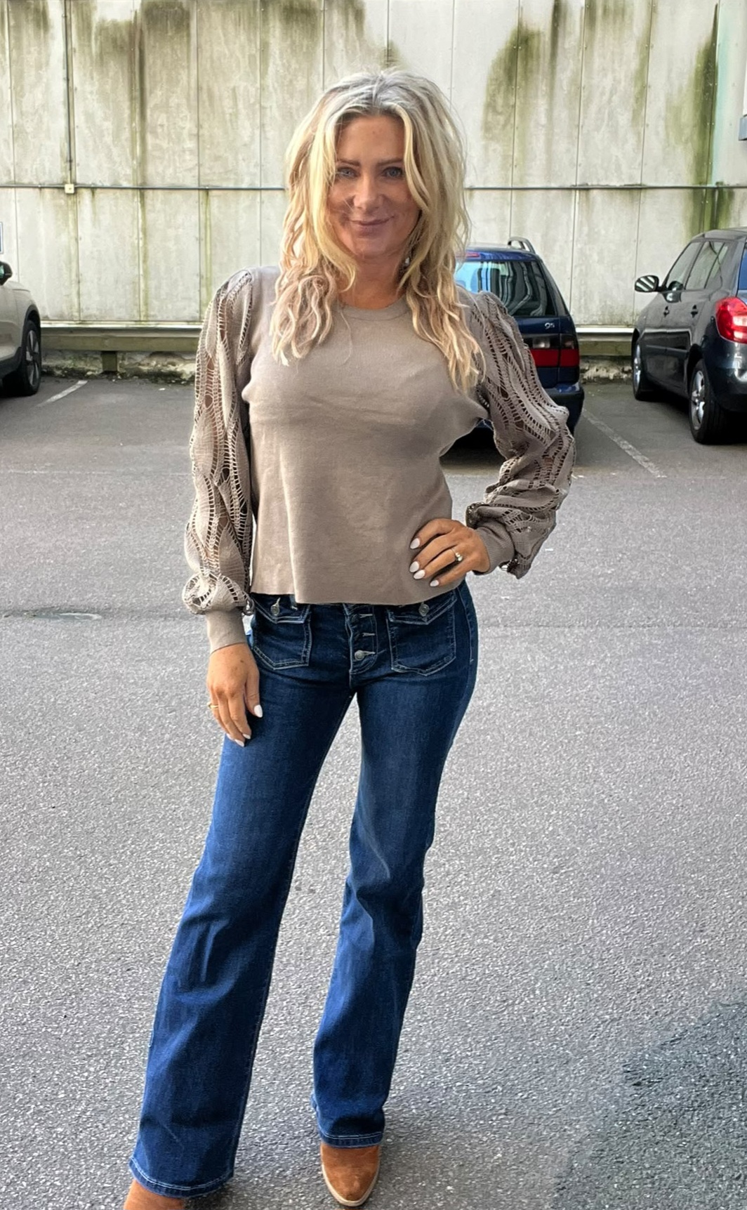 Blond kvinna står och poserar med ena handen på höften på en parkeringsplats. Hon bär beige topp med spetsiga ballongärmar, mörkblå bootcut-jeans och bruna skor; bilar och en betongvägg syns i bakgrunden.