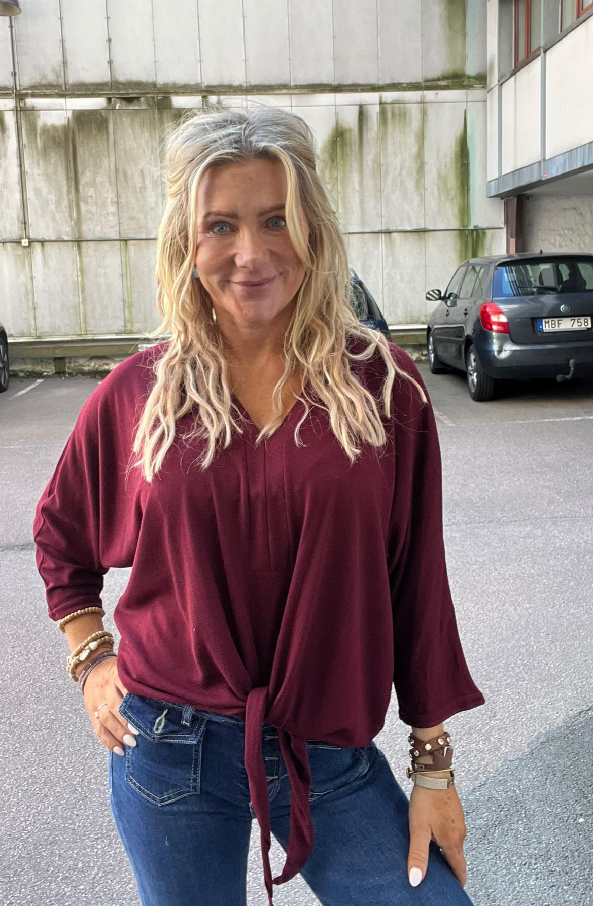 Leende kvinna med långt blont hår står med ena handen på höften och tittar mot kameran. Hon bär vinröd knytblus och jeans. Bakom syns parkering med bilar och en sliten betongvägg. Registreringsskylt: MBF 758.