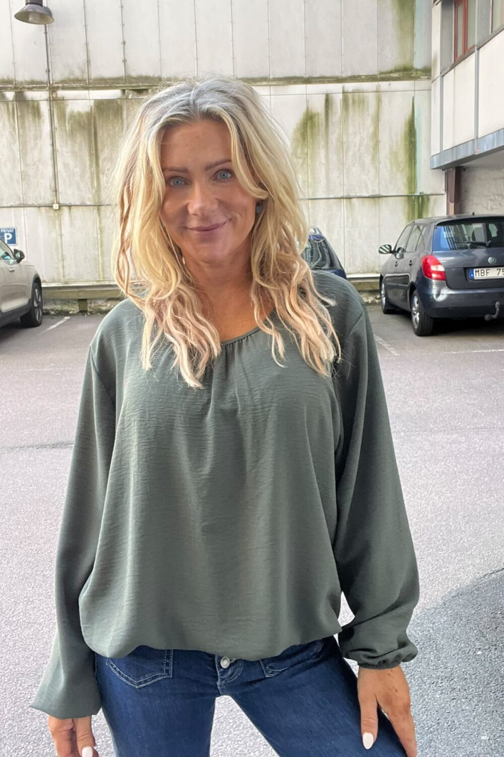 Kvinna med långt blont hår står och ler mot kameran, klädd i grön långärmad blus och blå jeans. Hon står på en parkeringsplats framför en betongvägg med skylten “PRIVAT P”.