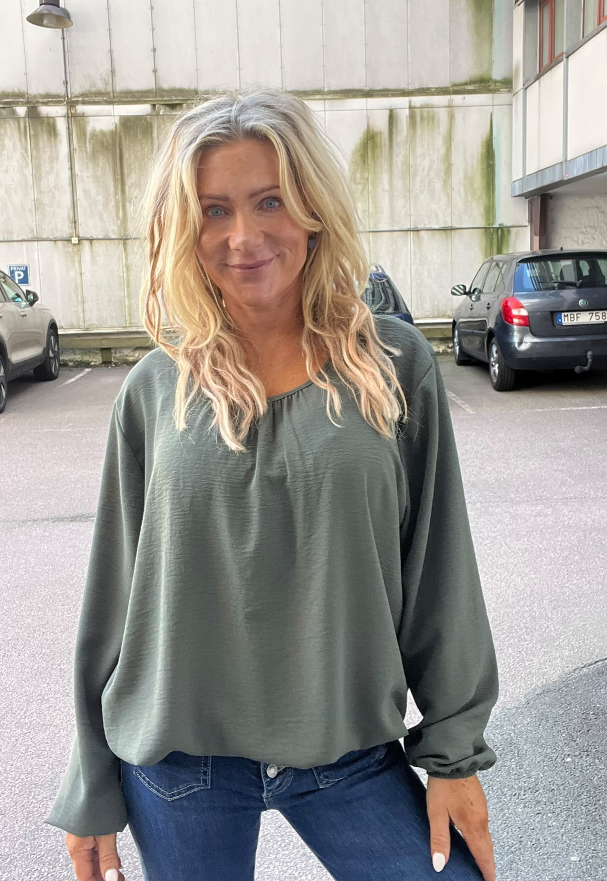 Kvinna med långt blont hår står och ler mot kameran, klädd i grön långärmad blus och blå jeans. Hon står på en parkeringsplats framför en betongvägg med skylten “PRIVAT P”.