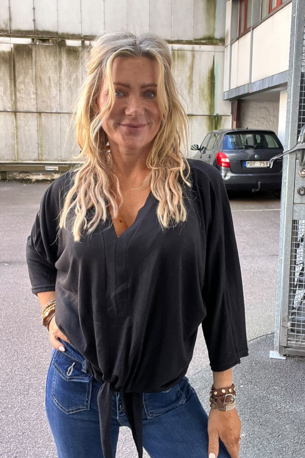 Blond kvinna i svart, knuten blus och blå jeans står och ler mot kameran med en hand i fickan. Hon står utomhus vid en industribyggnad; en parkerad bil syns i bakgrunden.