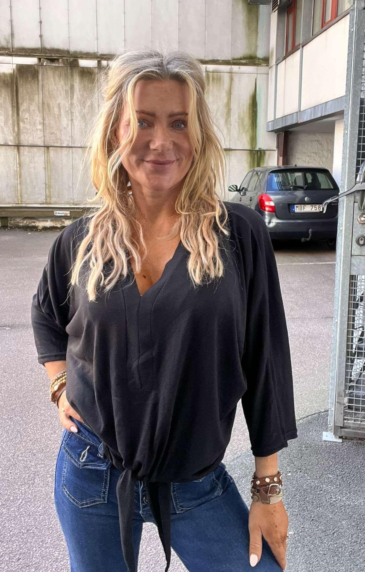 Blond kvinna i svart, knuten blus och blå jeans står och ler mot kameran med en hand i fickan. Hon står utomhus vid en industribyggnad; en parkerad bil syns i bakgrunden.
