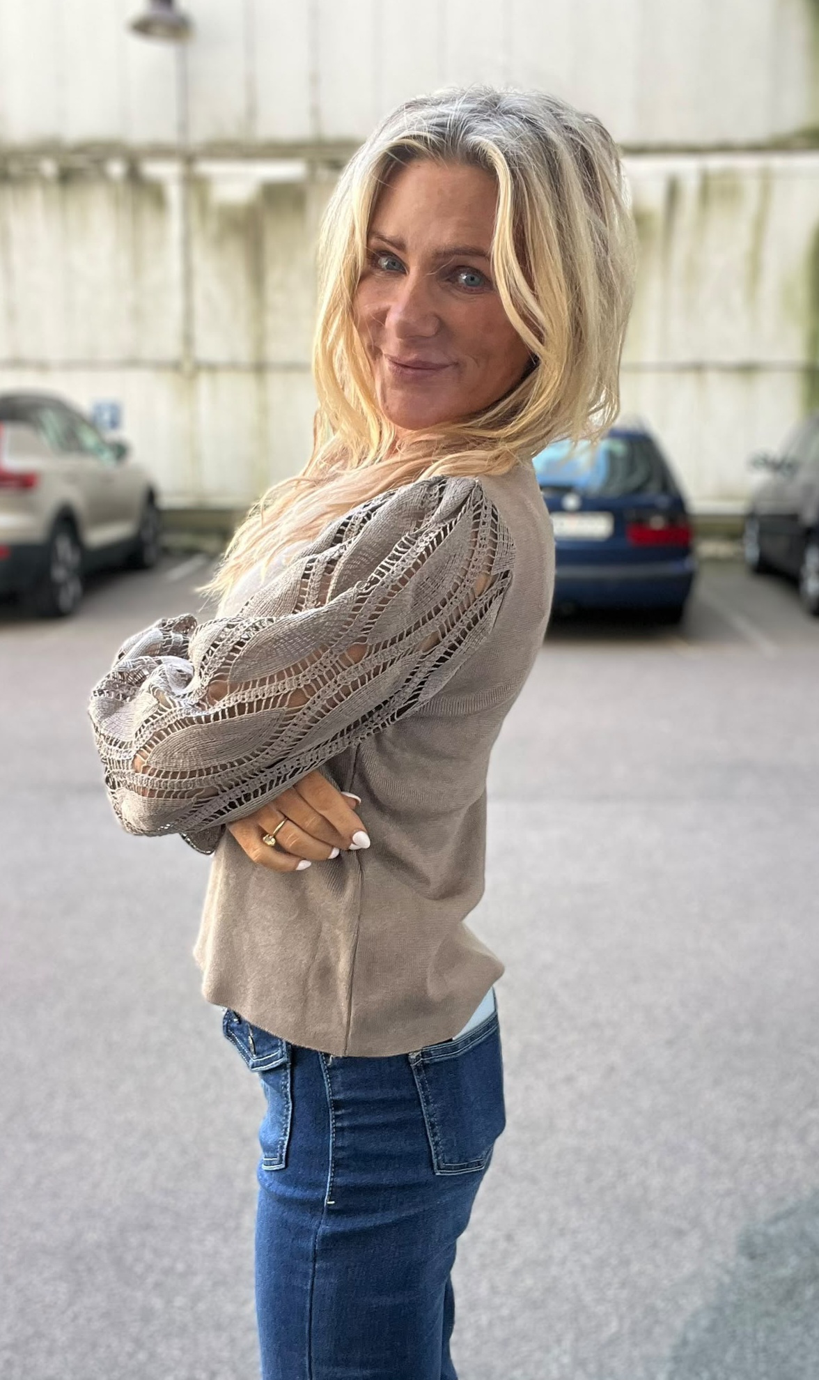 Blond kvinna står med armarna i kors och ler mot kameran på en parkeringsplats. Hon bär beige topp med genombrutna spetsärmar och jeans; suddiga bilar och en ljus vägg syns i bakgrunden.