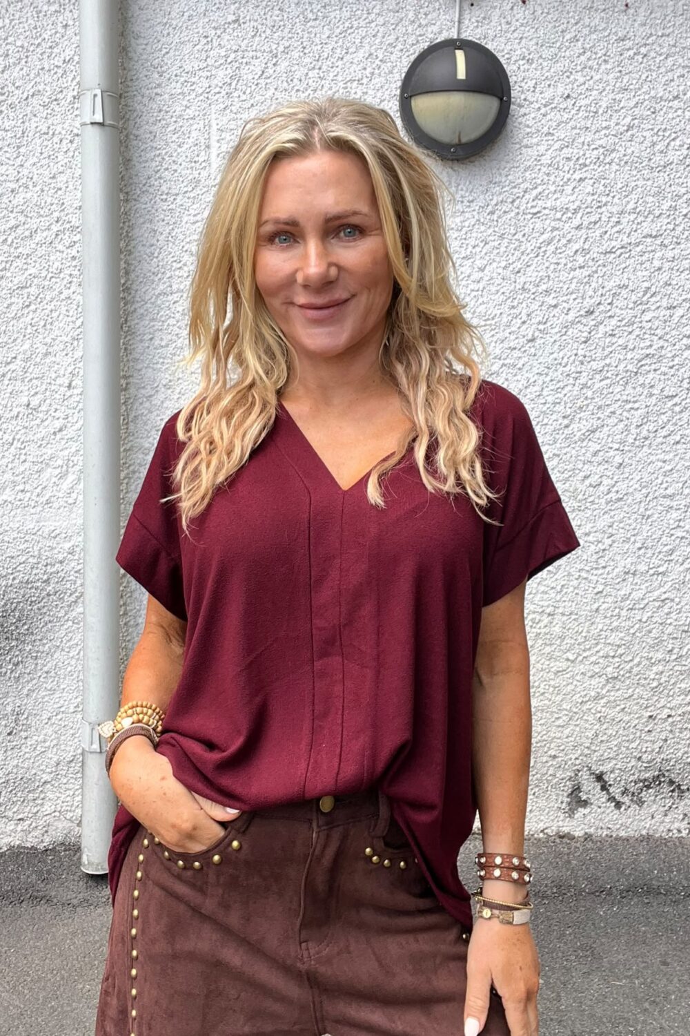 Blond kvinna står och ler mot kameran med ena handen i fickan. Hon bär vinröd V-ringad topp, bruna byxor med nitar och flera armband. Bakom henne syns en vit putsad vägg och en rund vägglampa.