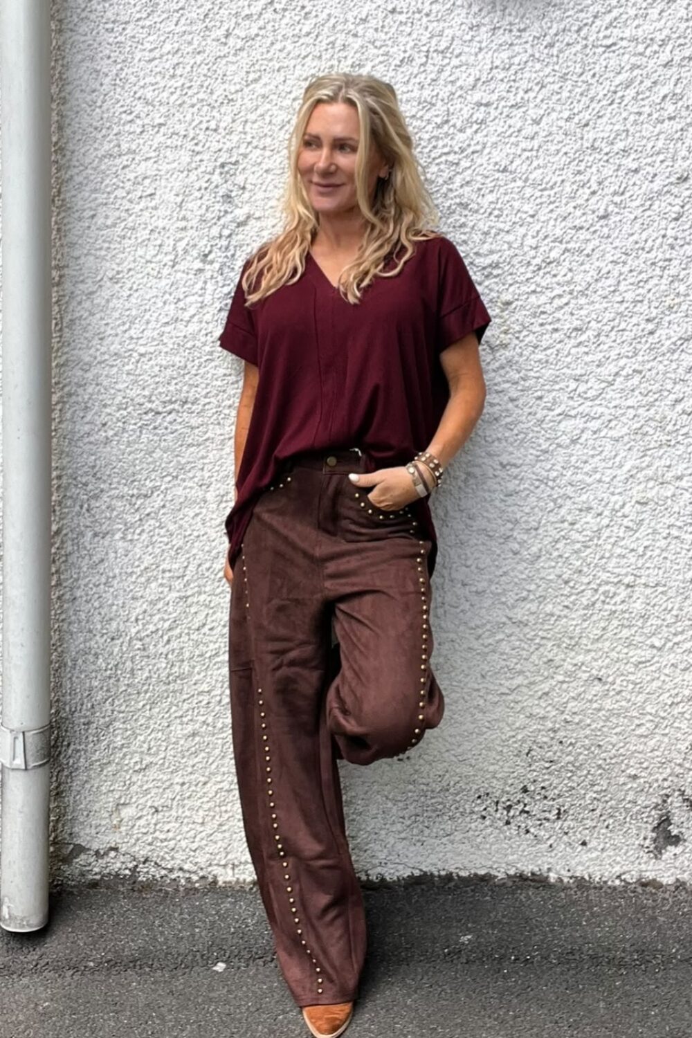 Kvinna med långt blont hår lutar sig mot en ljus, grovputsad vägg och tittar åt sidan. Hon har vinröd topp, bruna byxor med nitar, en hand i fickan och bruna skor; utomhus vid en vägglampa.