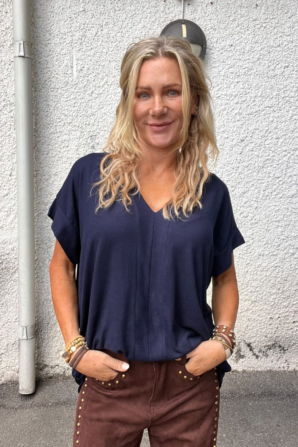 Leende kvinna med vågigt blont hår står utomhus mot en vit putsad vägg och ser in i kameran. Hon har händerna i fickorna, bär mörkblå topp och bruna byxor med nitar.