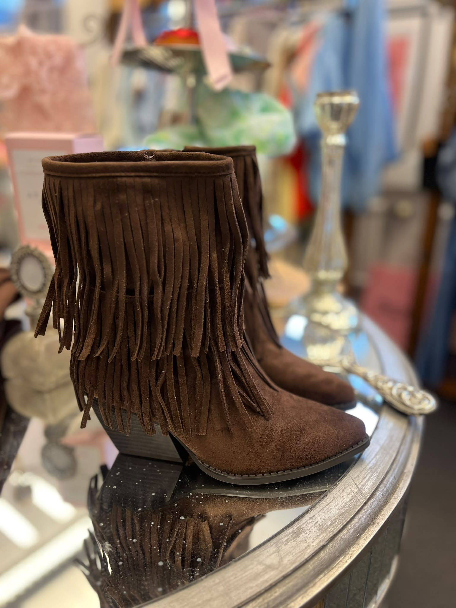 Bruna mockaboots med låg klack och långa fransar står uppställda på ett runt glasbord; fransarna hänger ner runt skaften. I bakgrunden syns suddiga butiksdetaljer som en glasljusstake och kläder.