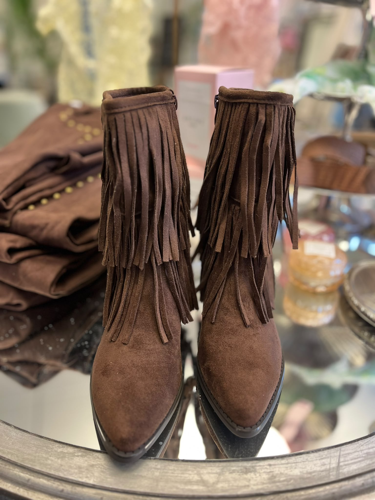 Bruna mockastövletter med spetsig tå står sida vid sida på ett bord, med flera lager långa fransar runt skaften. I bakgrunden syns en matchande brun väska och andra accessoarer i en butiksmiljö.