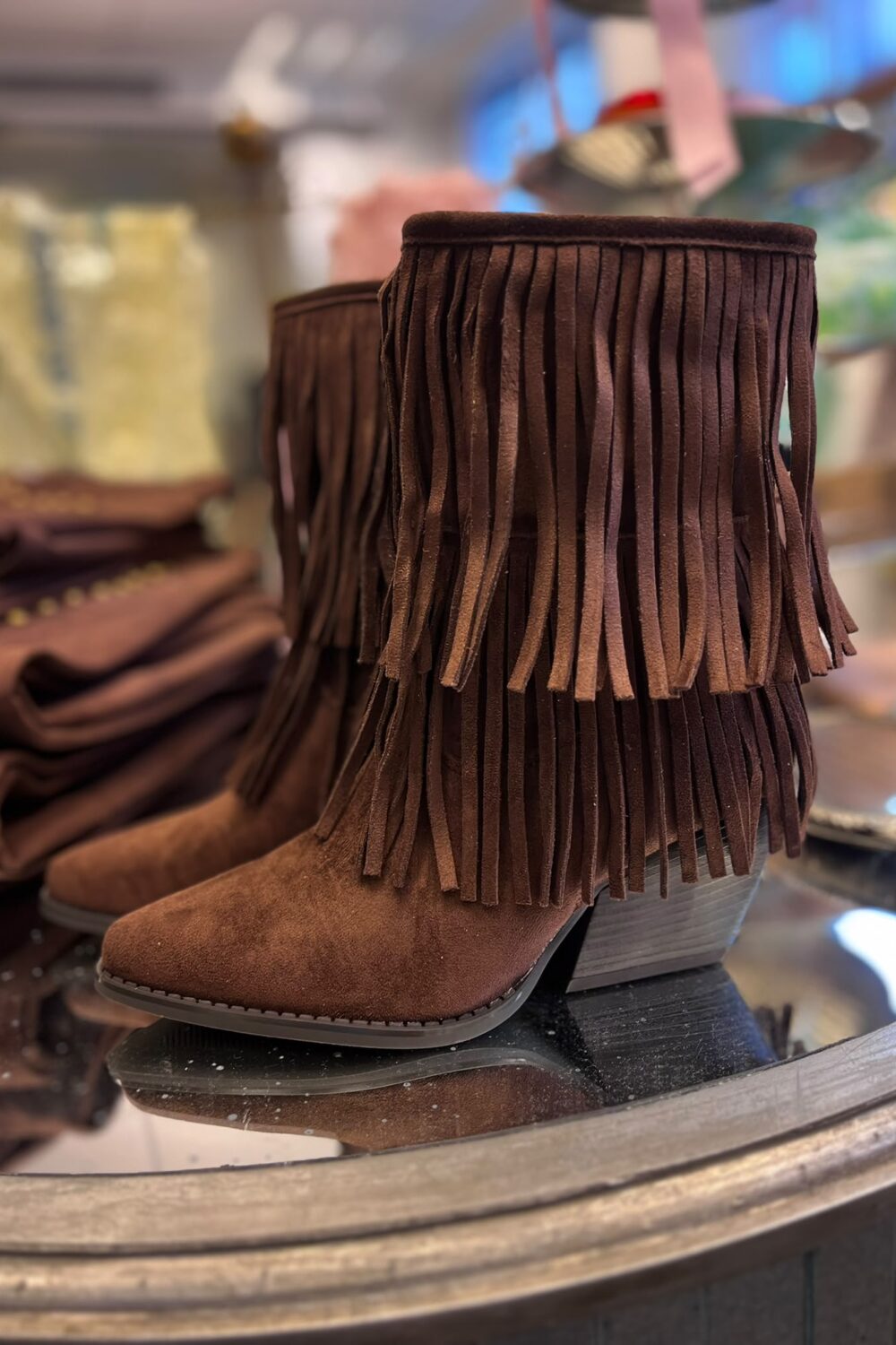 Brun mockastövel med kilklack och långa fransar står uppställd på en butikshylla; en andra stövel syns bakom. Bakgrunden är suddig med fler bruna skor och väskor, som i en skobutik.