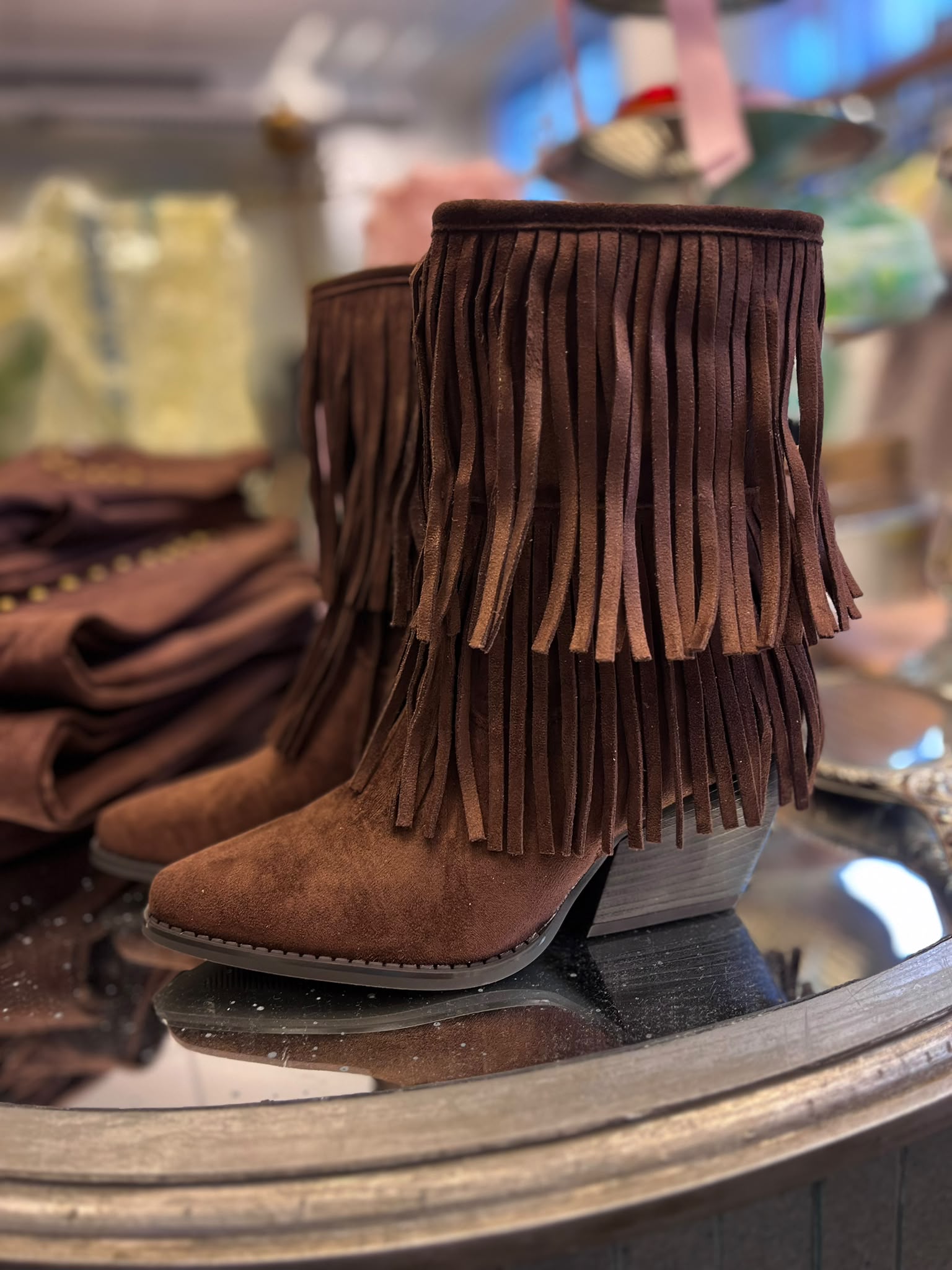 Brun mockastövel med kilklack och långa fransar står uppställd på en butikshylla; en andra stövel syns bakom. Bakgrunden är suddig med fler bruna skor och väskor, som i en skobutik.