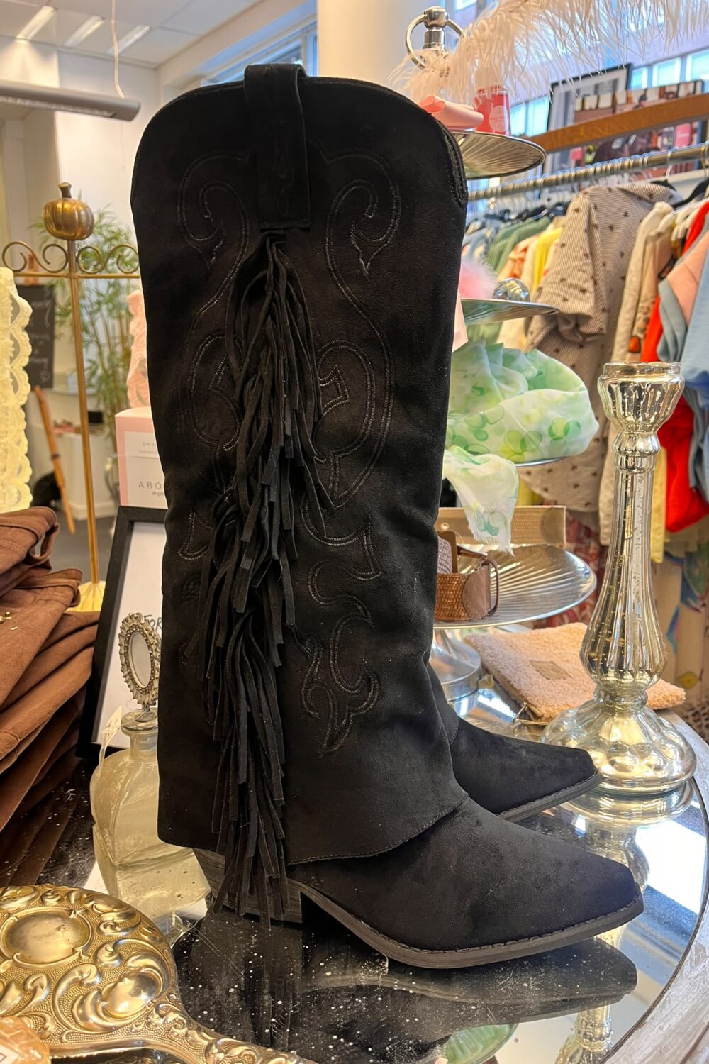 Svarta knähöga mockastövlar med broderade mönster och lång fransdetalj står uppställda på ett runt glasbord i en butik, omgivna av en silverfärgad ljusstake, dekorföremål och klädställning med färgglada plagg.