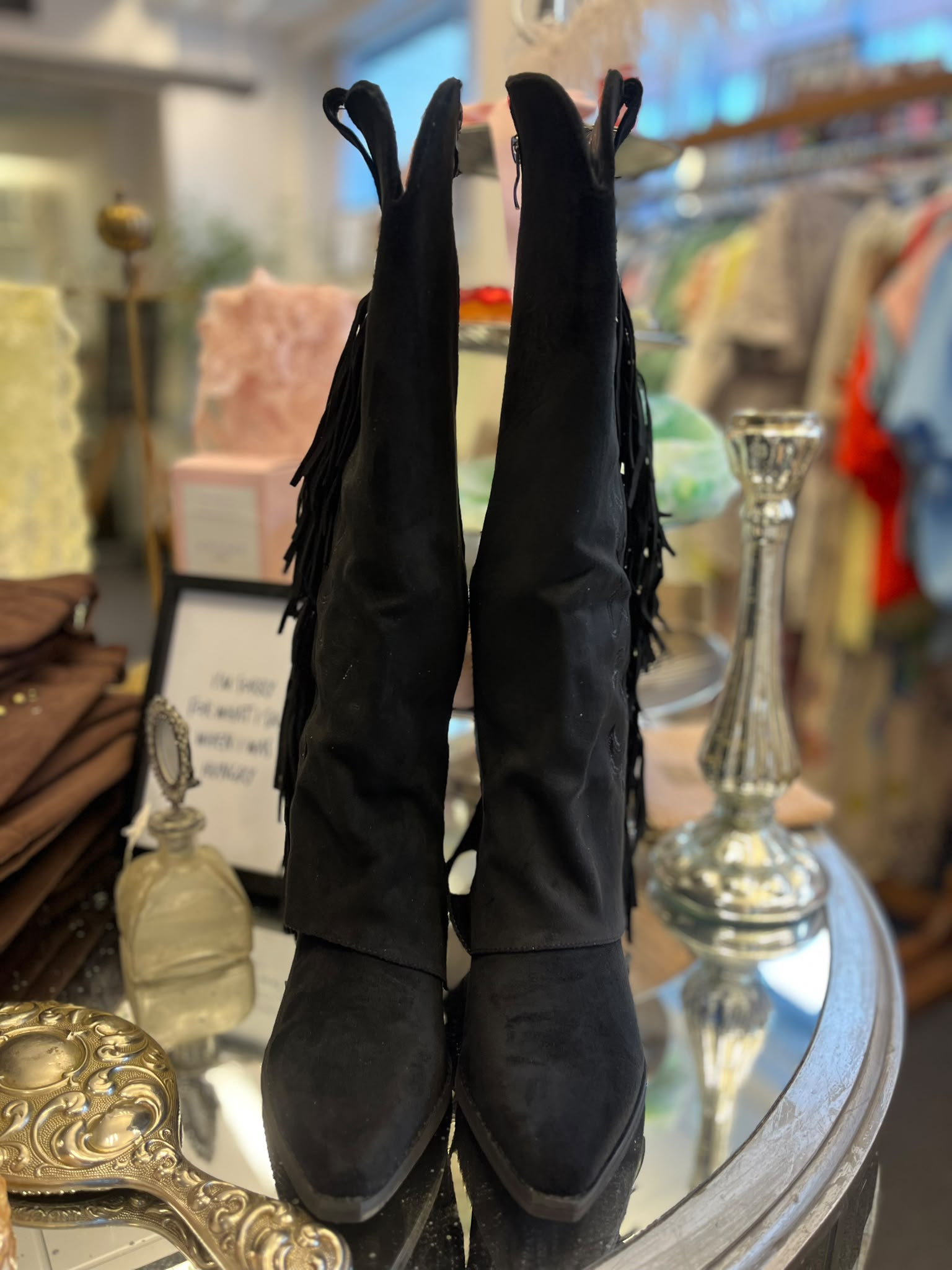 Ett par svarta knähöga stövlar med fransar står uppställda på ett runt glasbord. De är fotograferade i en butiksmiljö med suddiga kläder, lampor och dekorföremål i bakgrunden.