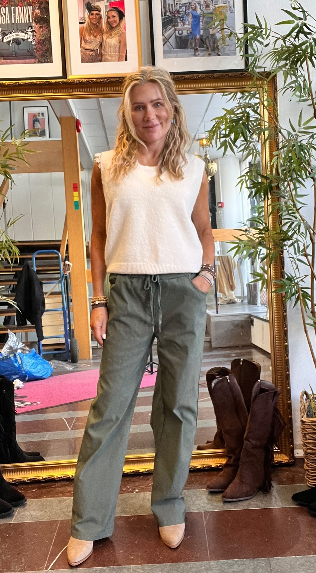 Blond kvinna står avslappnat med ena handen i fickan och ler mot kameran, klädd i vit ärmlös stickad topp, gröna byxor och beige skor. Bakom henne syns en stor guldspegel, växter och ett par stövlar.
