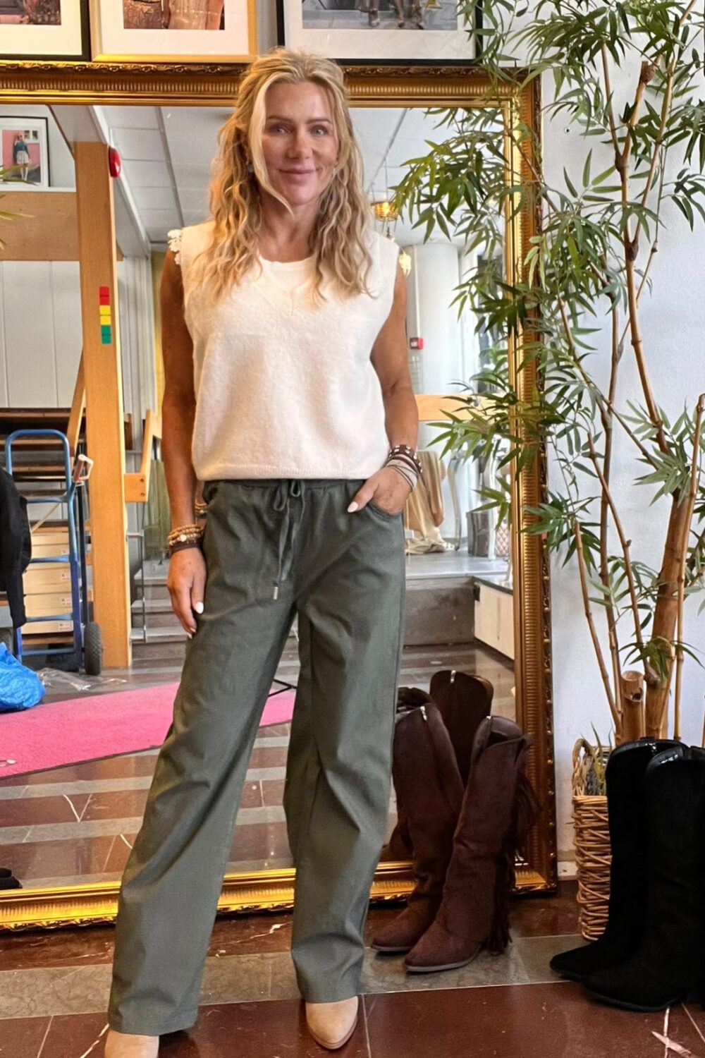 Blond kvinna står och ler med ena handen i fickan, klädd i vit ärmlös topp, gröna byxor och beige klackskor. Hon poserar framför en stor guldramsspegel i en butik med boots och växt bredvid.