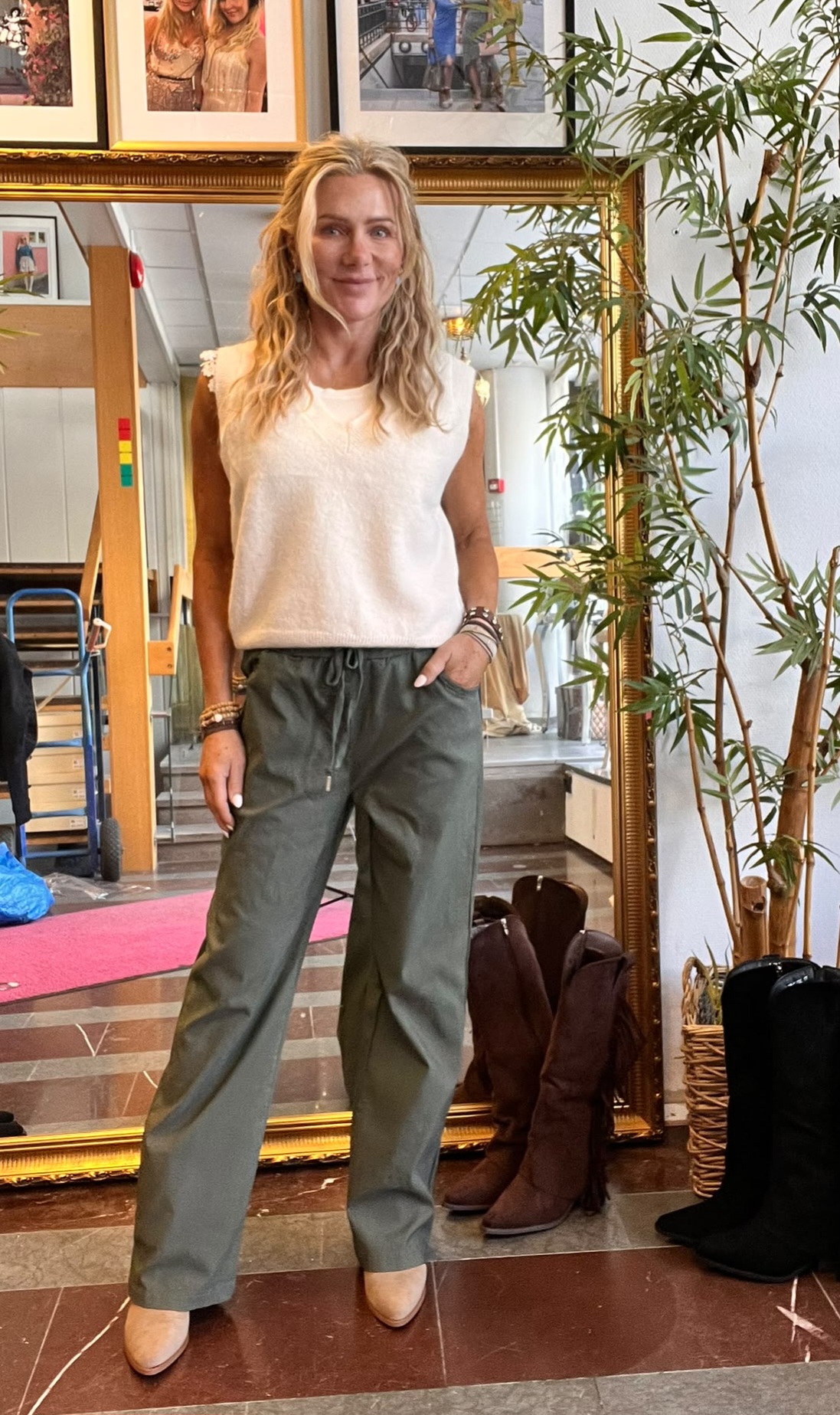 Blond kvinna står och ler med ena handen i fickan, klädd i vit ärmlös topp, gröna byxor och beige klackskor. Hon poserar framför en stor guldramsspegel i en butik med boots och växt bredvid.