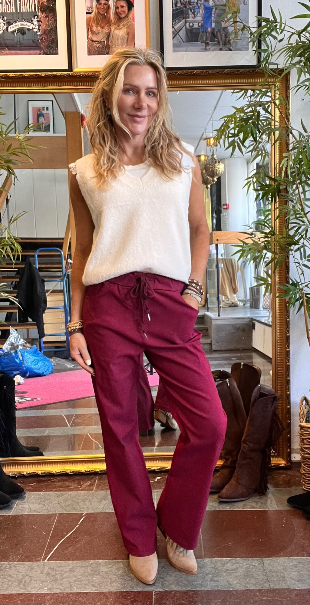 Blond kvinna poserar med ena handen i fickan framför en stor guldramad spegel, klädd i vit stickad topp och vinröda byxor med beige skor. Inomhusmiljö med växter, tavlor och skor på golvet.
