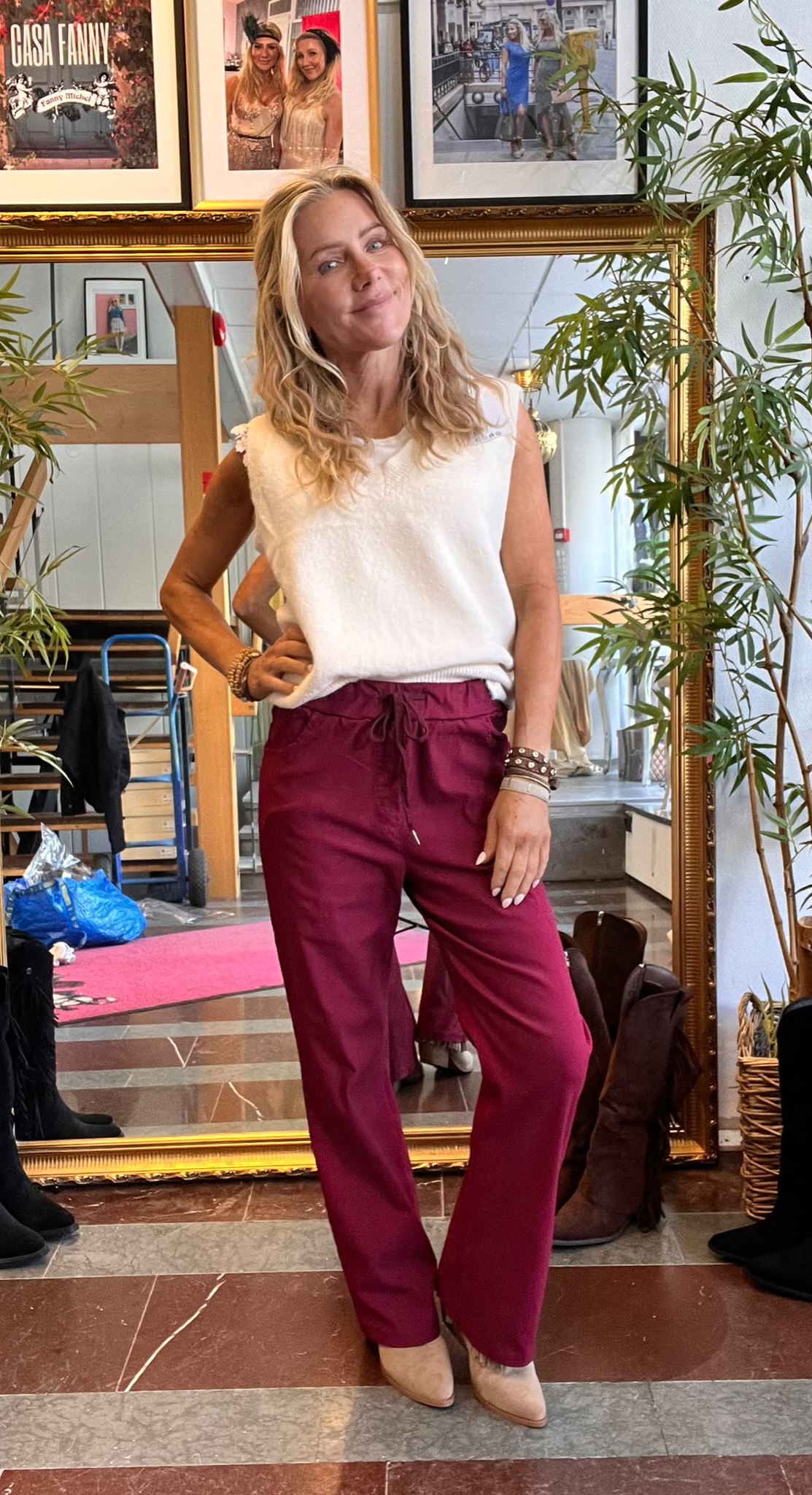 Blond kvinna poserar med handen på höften framför en stor guldramsspegel, klädd i vit topp och vinröda byxor. I bakgrunden syns växter, skor och inramade foton med texten ”CASA FANNY”.