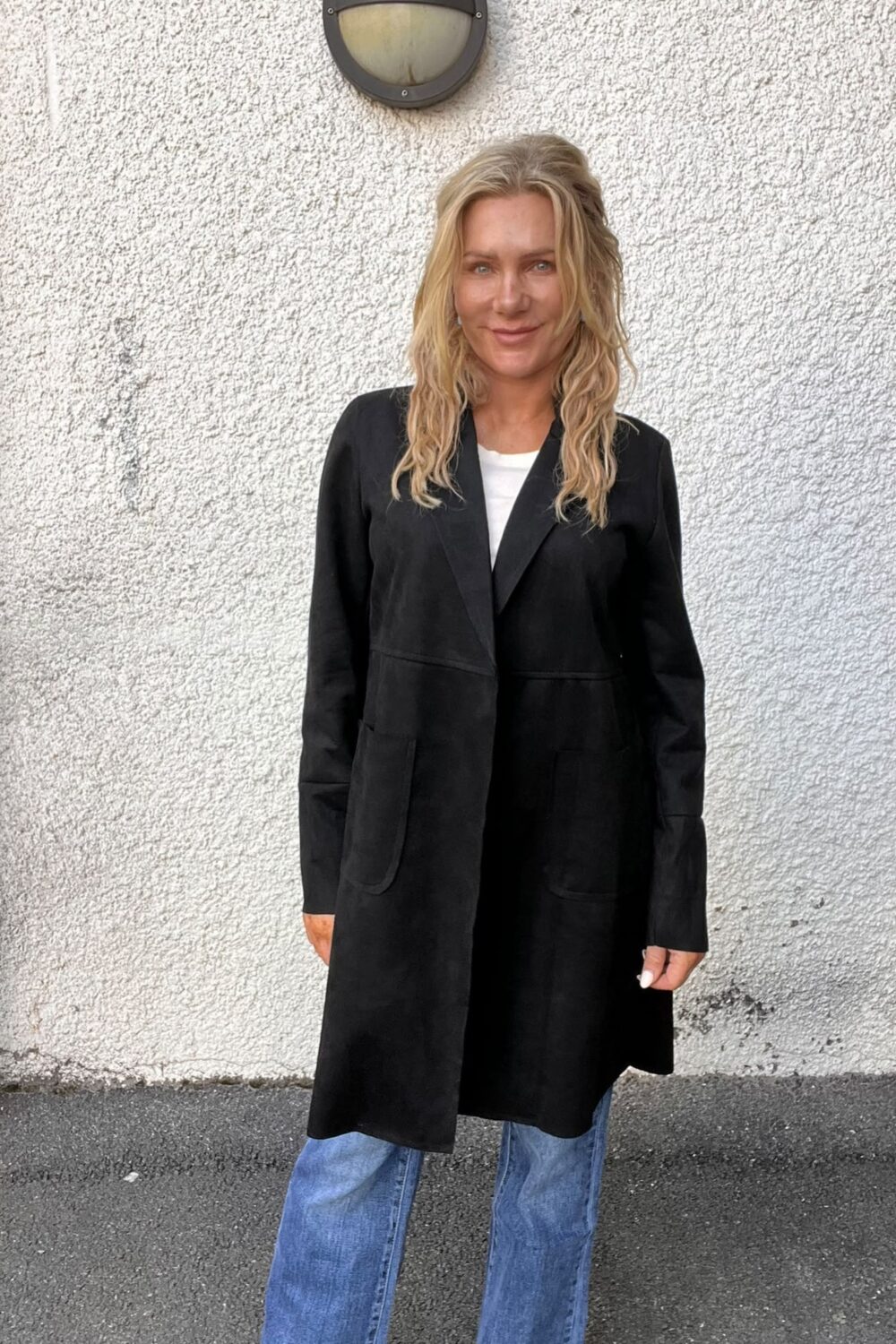 Blond kvinna står och ler mot kameran utomhus, framför en vit, grov putsad vägg med rund vägglampa ovanför. Hon bär lång svart kappa över vit topp och vida blå jeans.