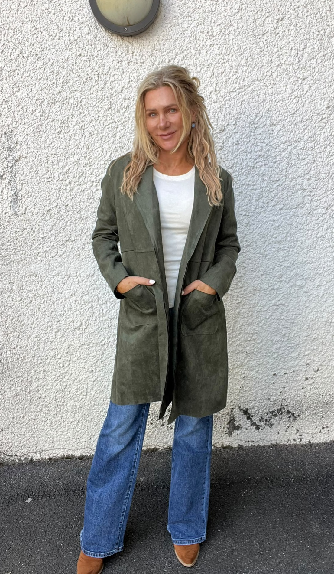 Kvinna står och ler mot kameran med händerna i fickorna, framför en vit putsad vägg med rund vägglampa. Hon bär olivgrön lång jacka, vit topp, utsvängda blå jeans och bruna skor.