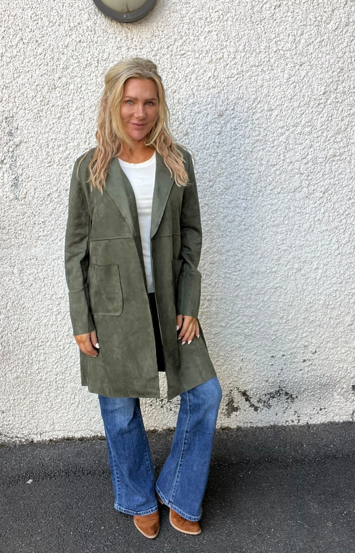 Kvinna med långt blont hår står och ler mot kameran framför en vit, grovputsad vägg utomhus. Hon bär en olivgrön lång kappa över vit tröja, blå bootcut-jeans och bruna skor.