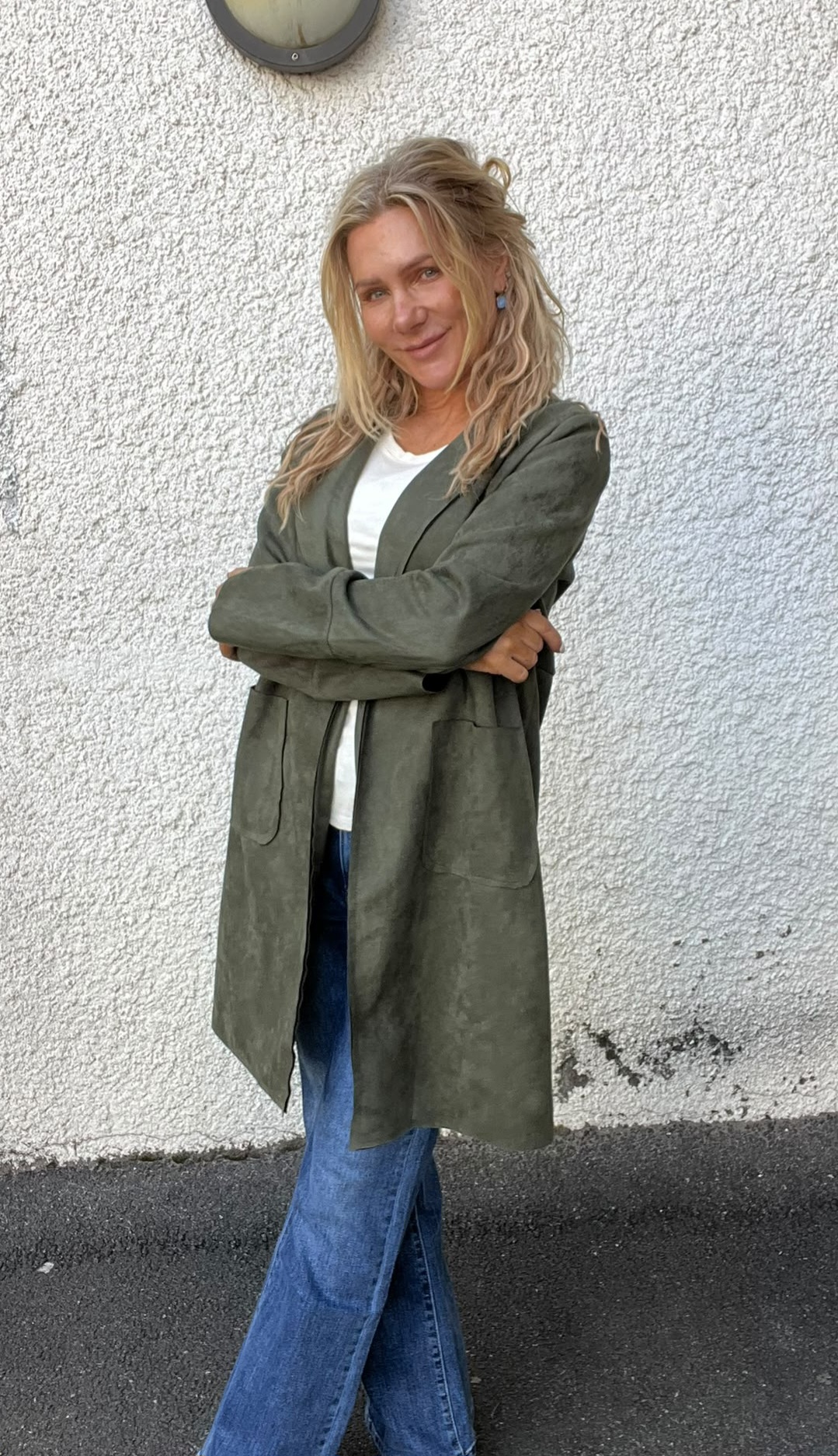 Kvinna med långt blont hår står med armarna i kors och ler mot kameran, klädd i olivgrön lång jacka, vit topp och vida jeans, framför en vit putsad vägg med en rund vägglampa ovanför.